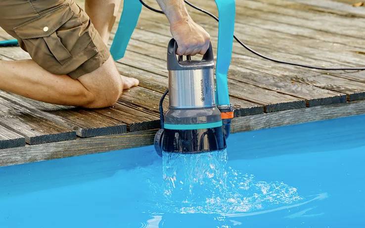 Person pumpt mit einer elektrischen Pumpe Wasser aus einem blauen Pool. Die Pumpe hat einen Metall- und Kunststoffkörper.