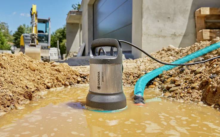 Eine Tauchpumpe pumpt Wasser aus einer Pfütze auf einer Baustelle ab. Ein Bagger ist unscharf im Hintergrund zu sehen.