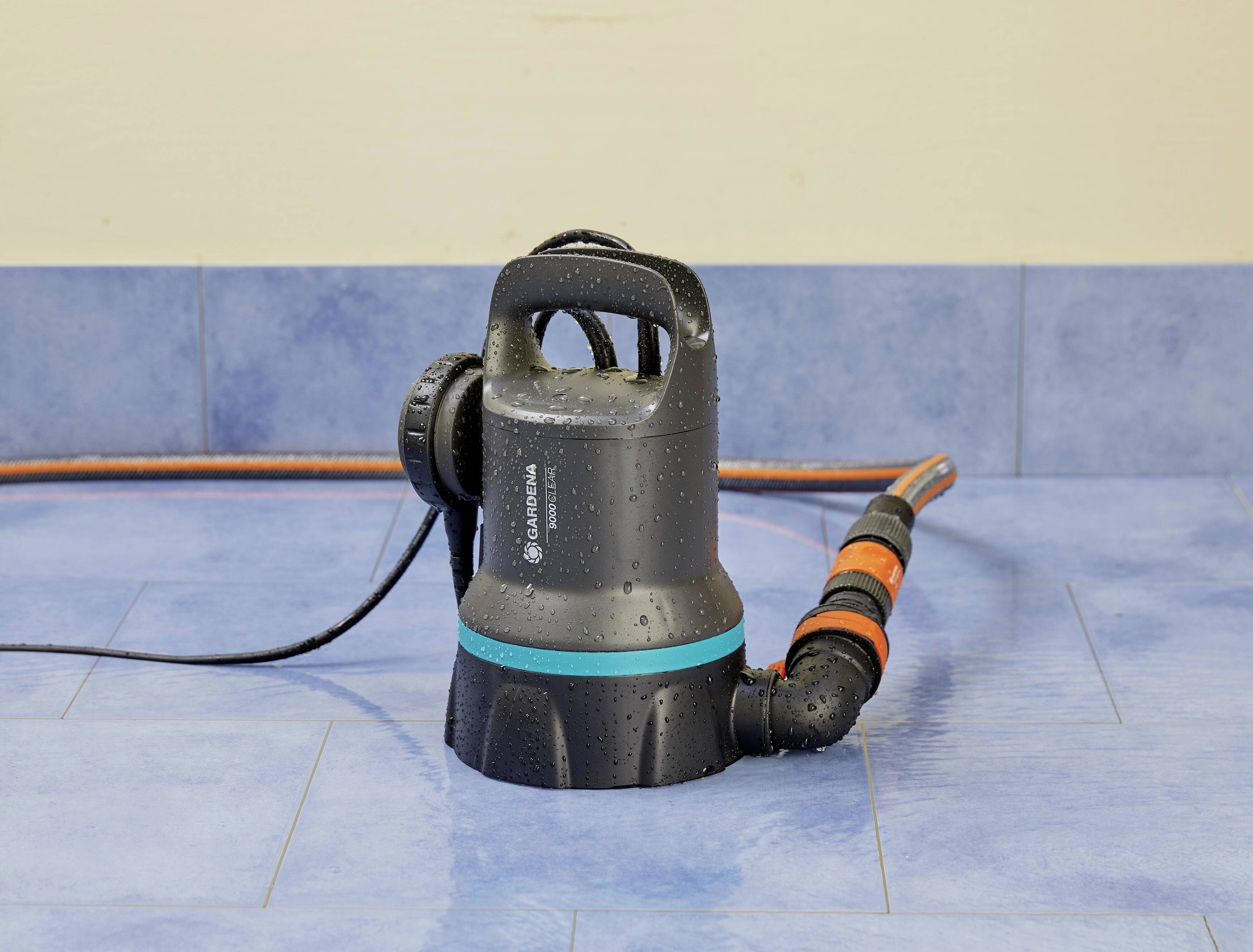 Eine schwarze Wasserpumpe steht auf blauem Bodenfliesen, mit Schläuchen, die daran befestigt sind. Die Pumpe ist feucht von Wasser.