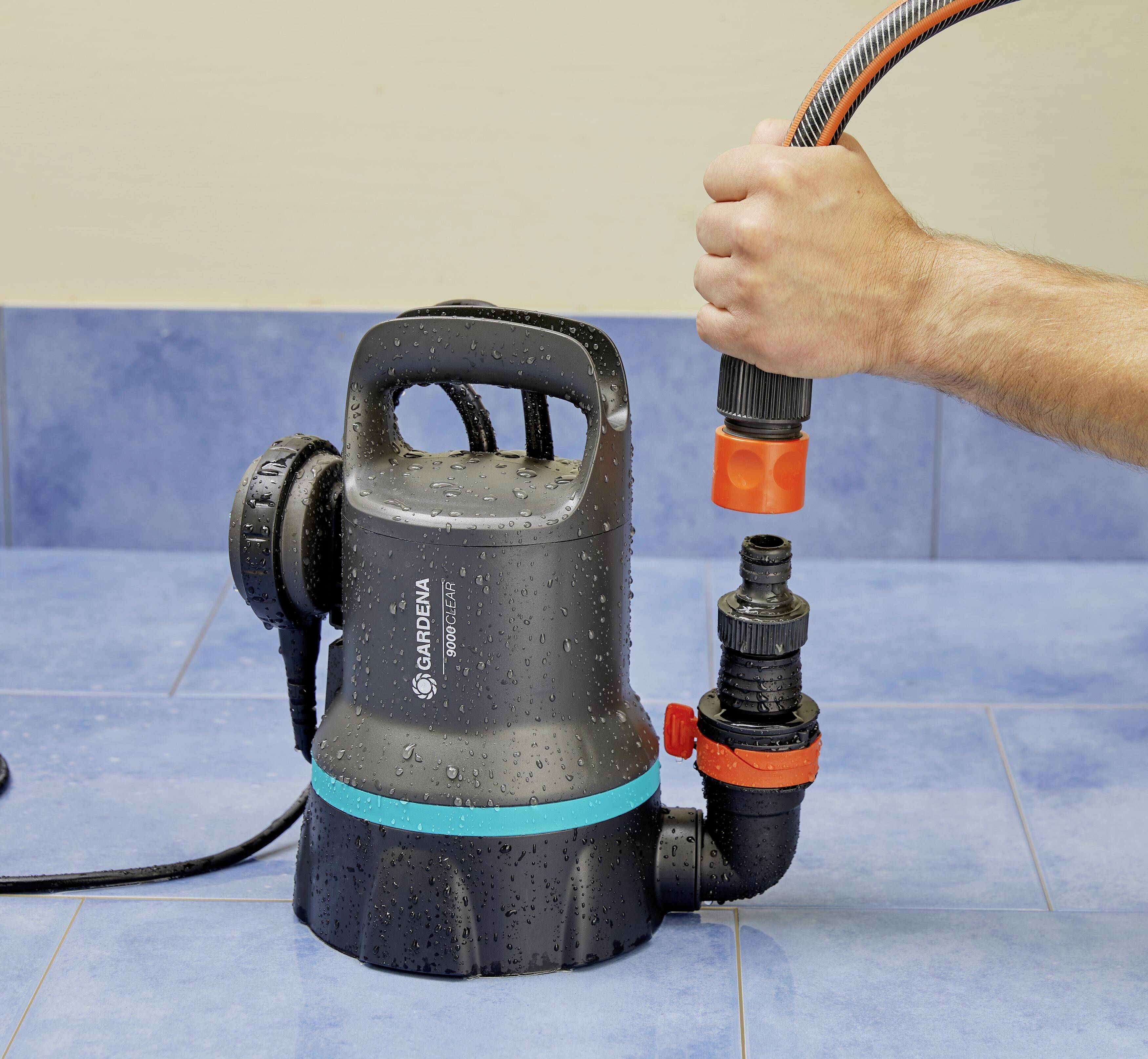 Eine Hand schließt einen Schlauch an eine schwarze Wasserpumpe an, die auf einem blauen Boden steht. Die Pumpe ist feucht vom Wasser.