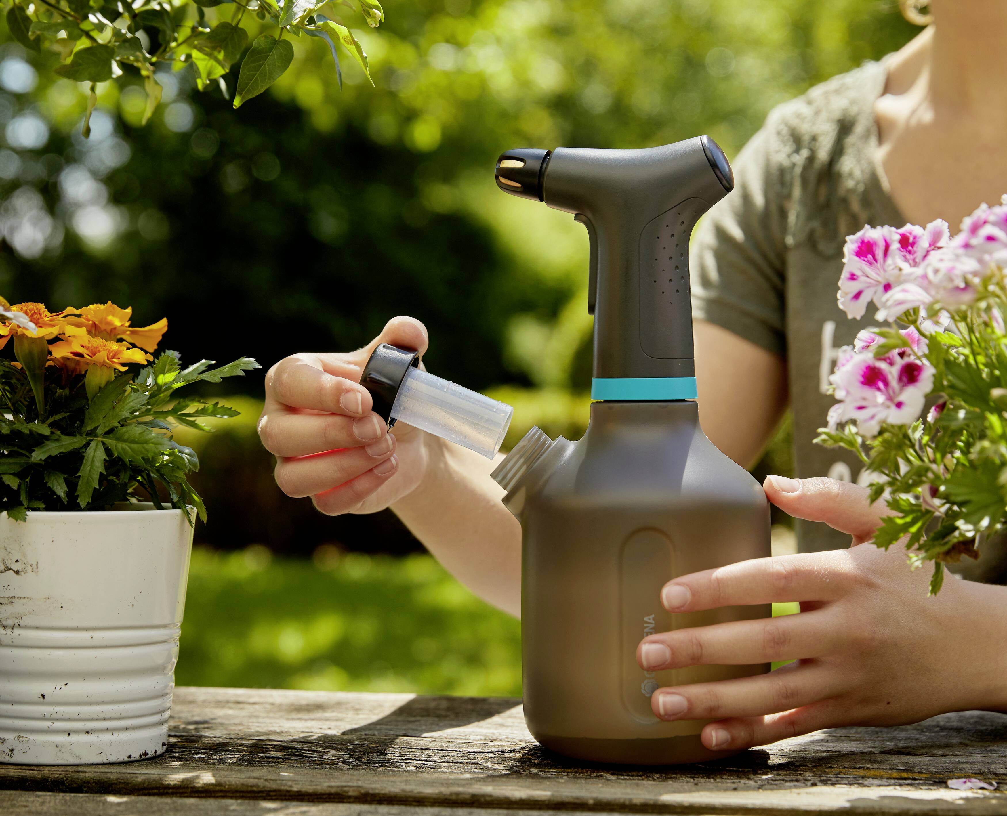 Eine Person befüllt eine graue Sprühflasche mit Flüssigkeit, umgeben von blühenden Pflanzen in weißen Töpfen auf einem Holztisch im Garten.