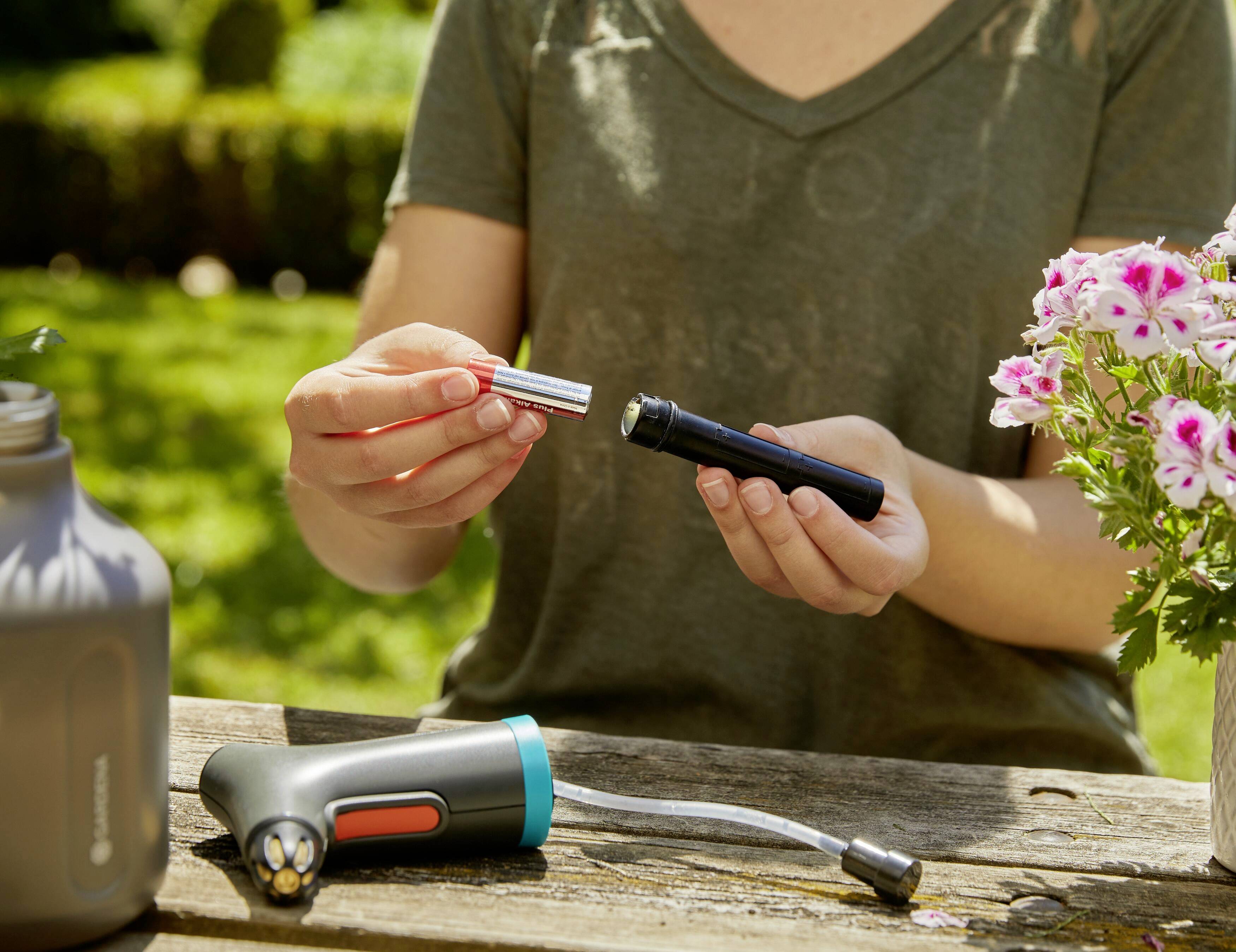 Eine Person sitzt im Freien und hält ein Batteriefach eines tragbaren Geräts. Blumen und ein anderes Gerät sind auf dem Tisch zu sehen.
