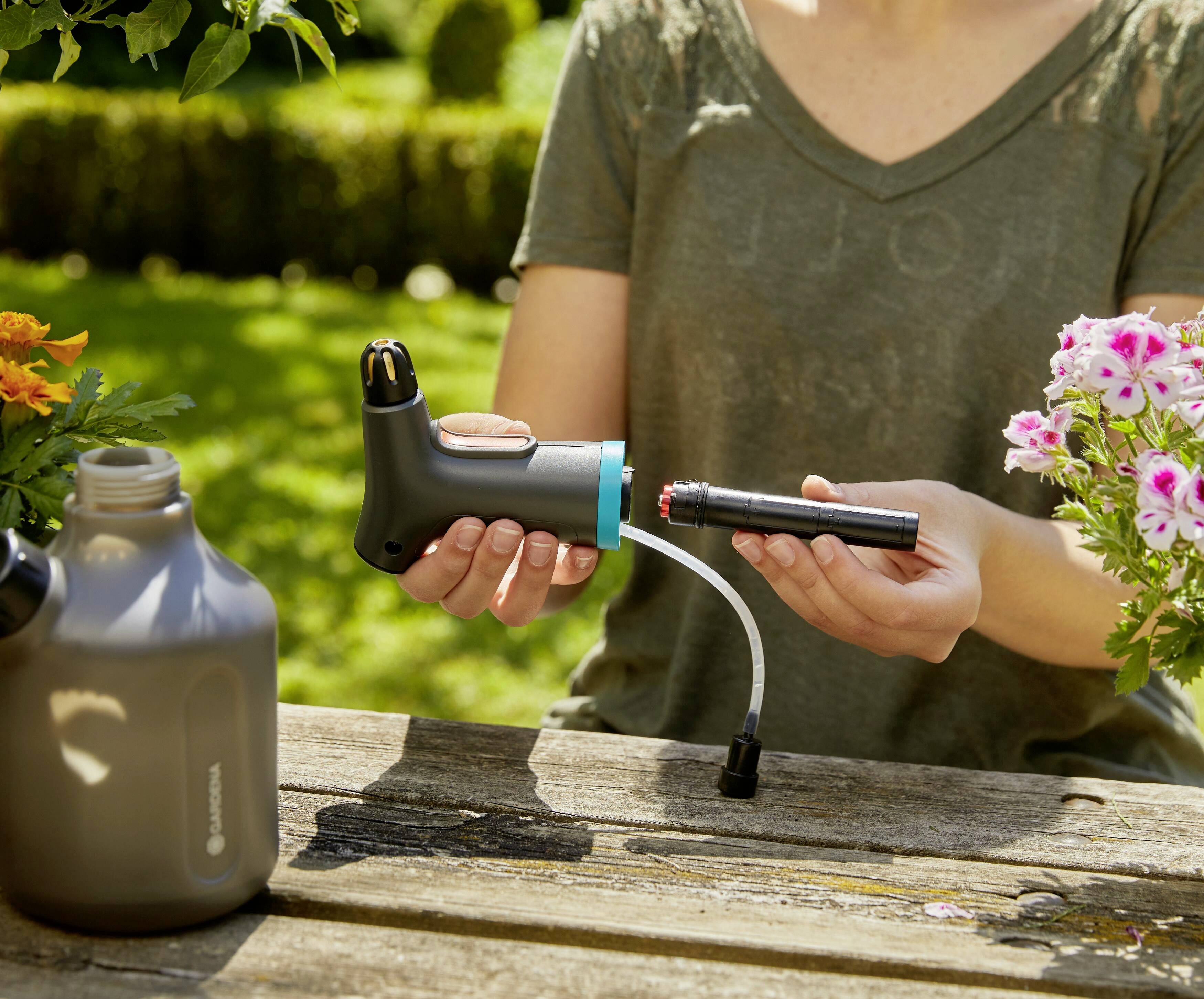Eine Person zeigt, wie ein Bewässerungsgerät zusammengebaut wird. Ein Tisch im Freien mit Blumen und einer Gießkanne ist zu sehen.