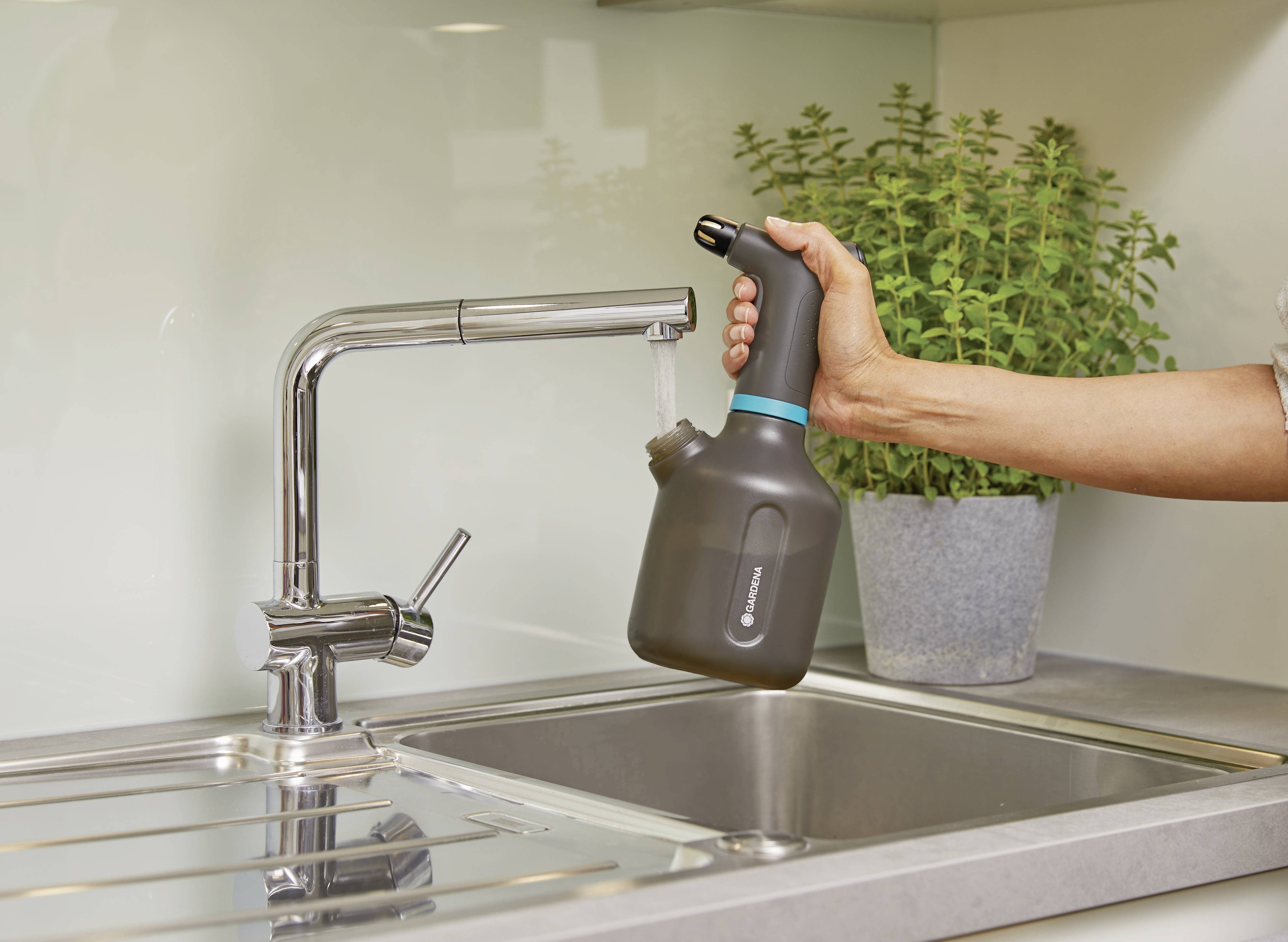 Eine Person hält eine Gießkanne unter einen Wasserhahn in einer Küche. Im Hintergrund steht eine Topfpflanze.