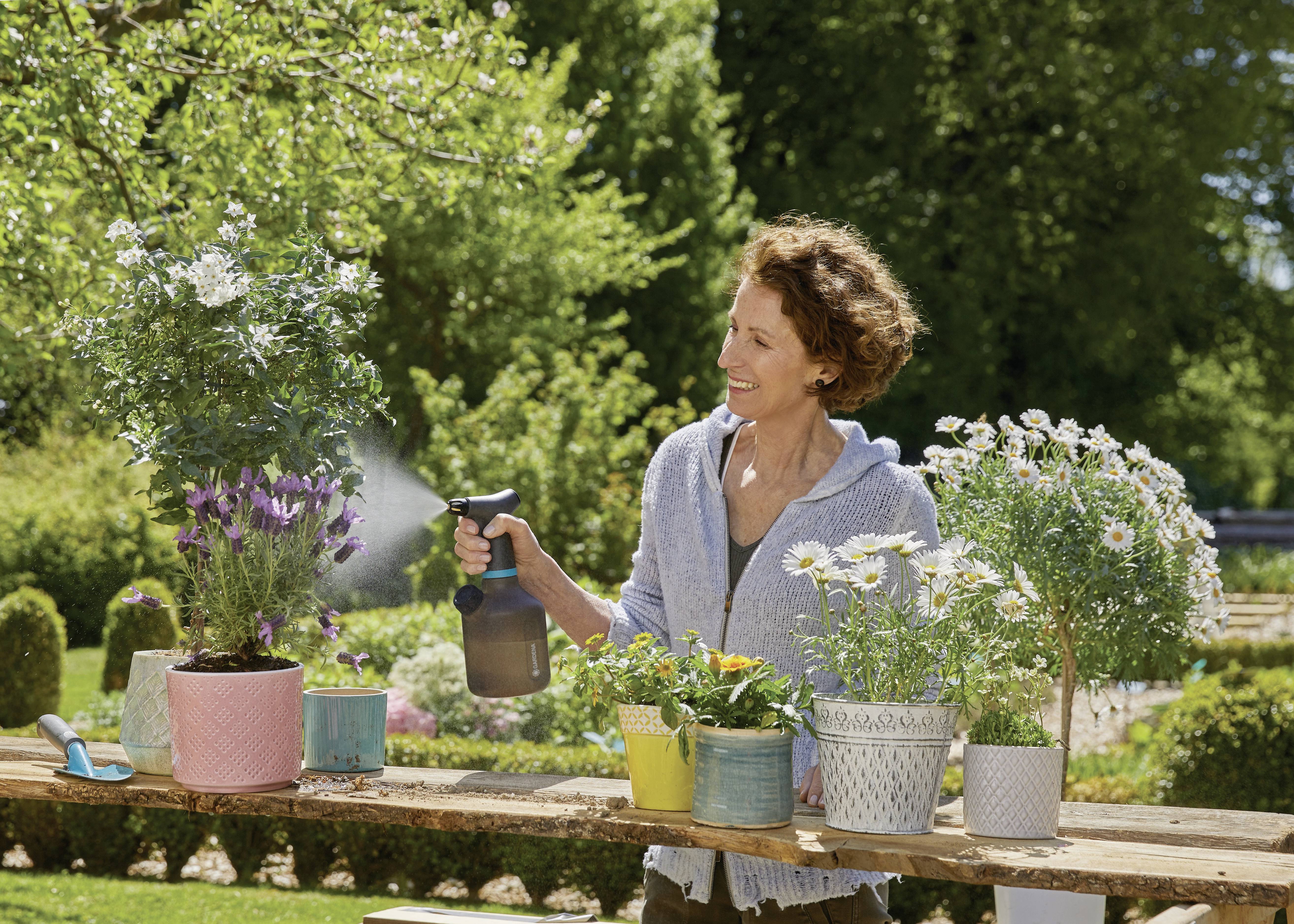 Eine lächelnde Frau gießt Blumen mit einer Sprühflasche im Garten. Verschiedene Topfpflanzen stehen auf einem Holztisch im Freien.