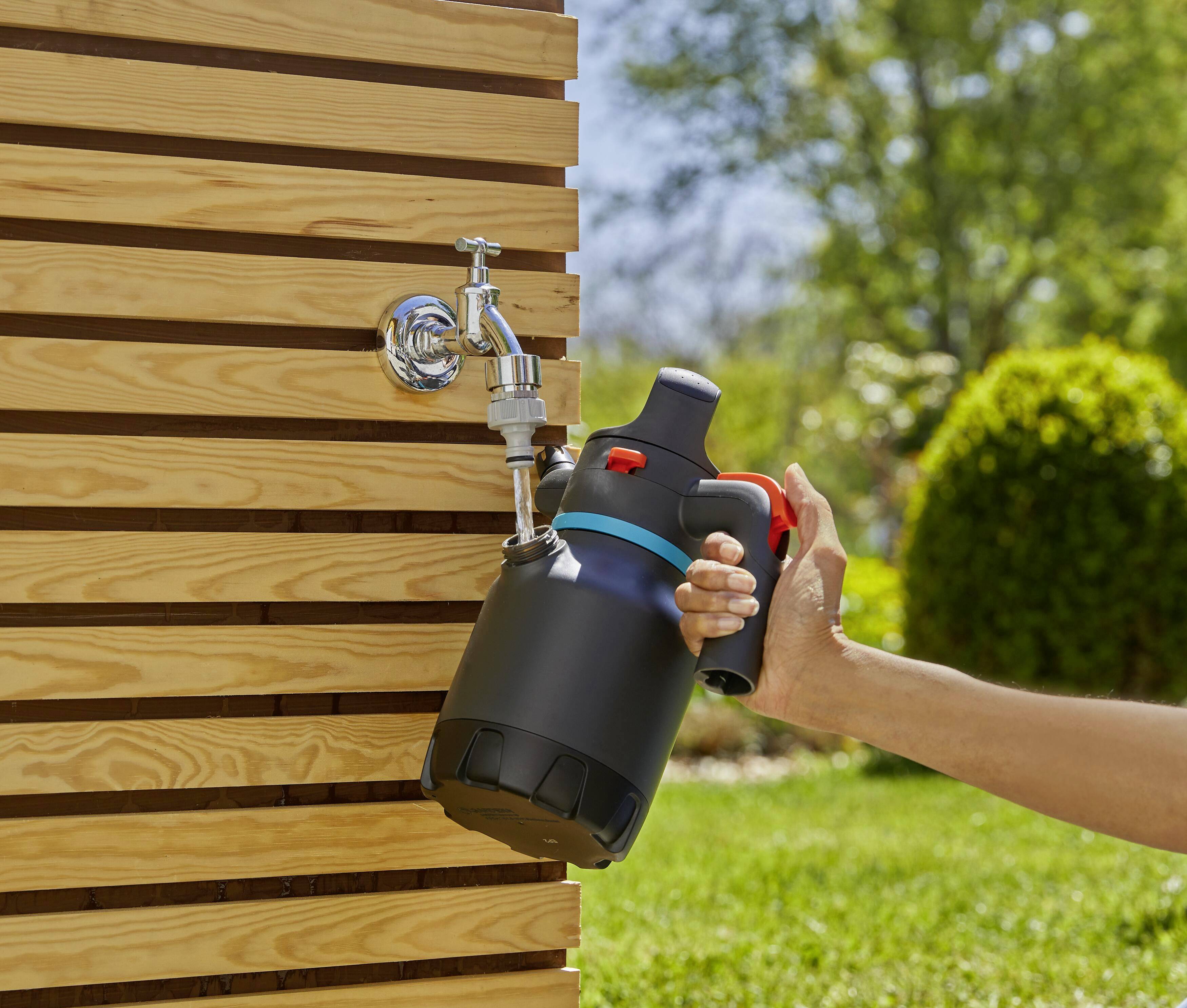 Eine Person füllt einen schwarzen Garten-Drucksprüher mit Wasser an einem Außenhahn, umgeben von einer grünen Wiese und Sonne.
