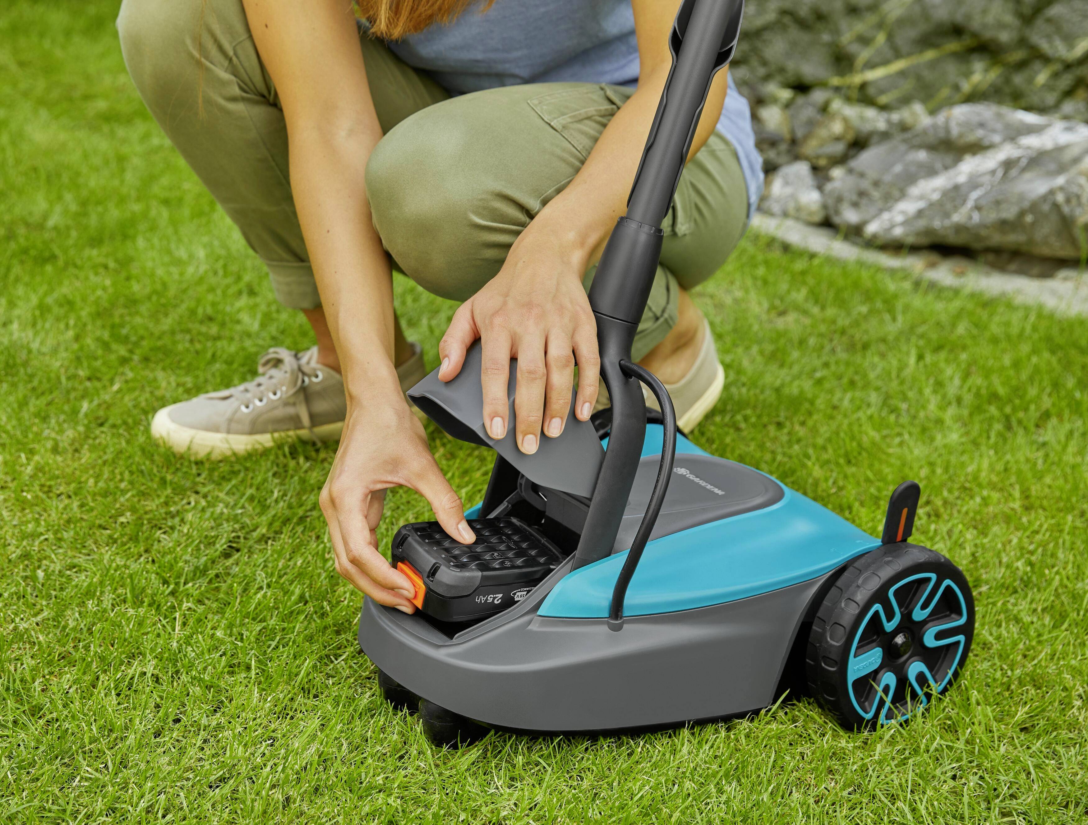 Eine Person ersetzt die Batterie eines Rasenmähers. Der Rasenmäher ist blau und schwarz, und die Person trägt lässige Kleidung.