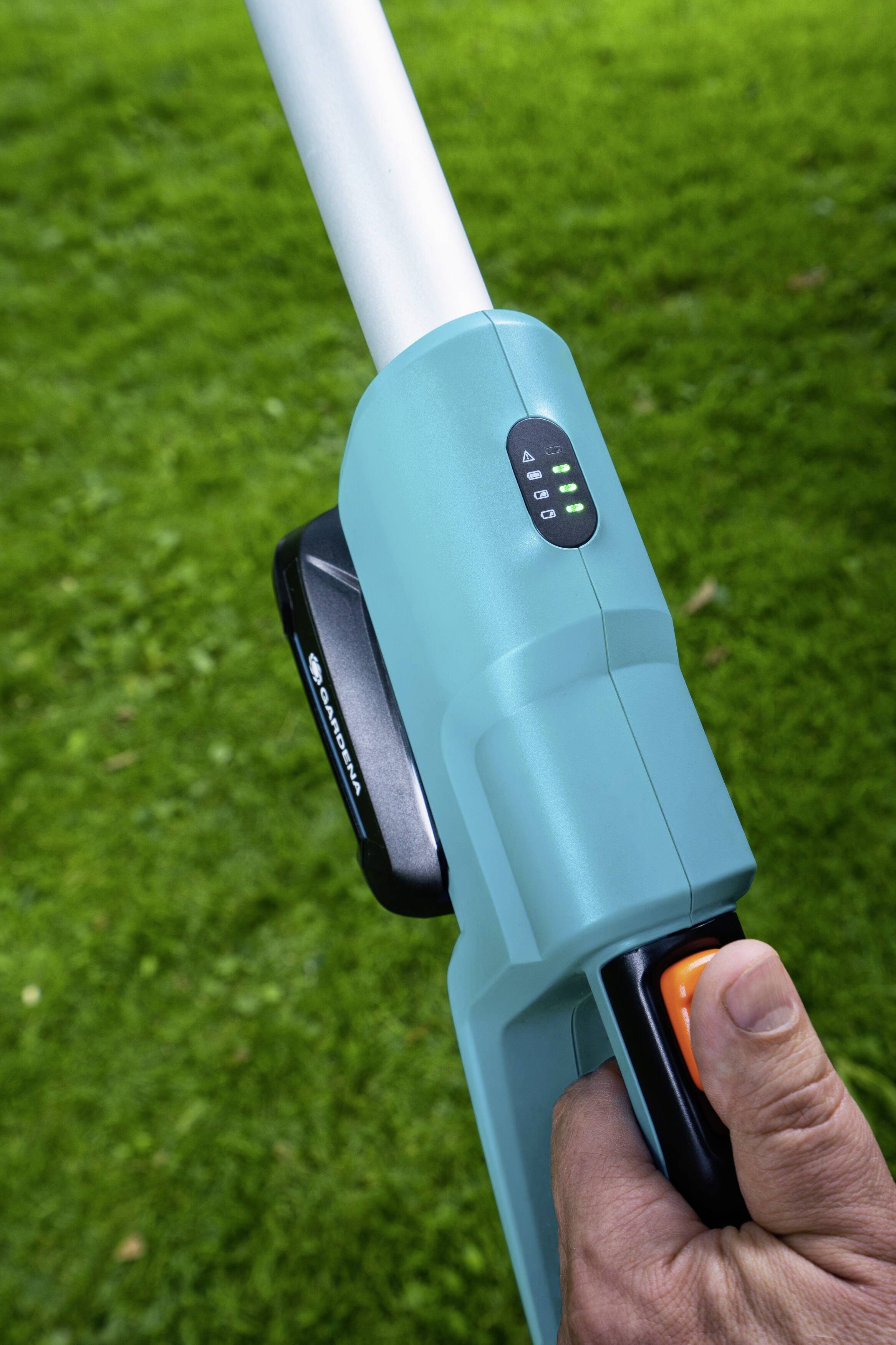 Person hält einen elektrischen Rasentrimmer, Akku und Ladestandsanzeige erkennbar. Hintergrund ist grüner Rasen.