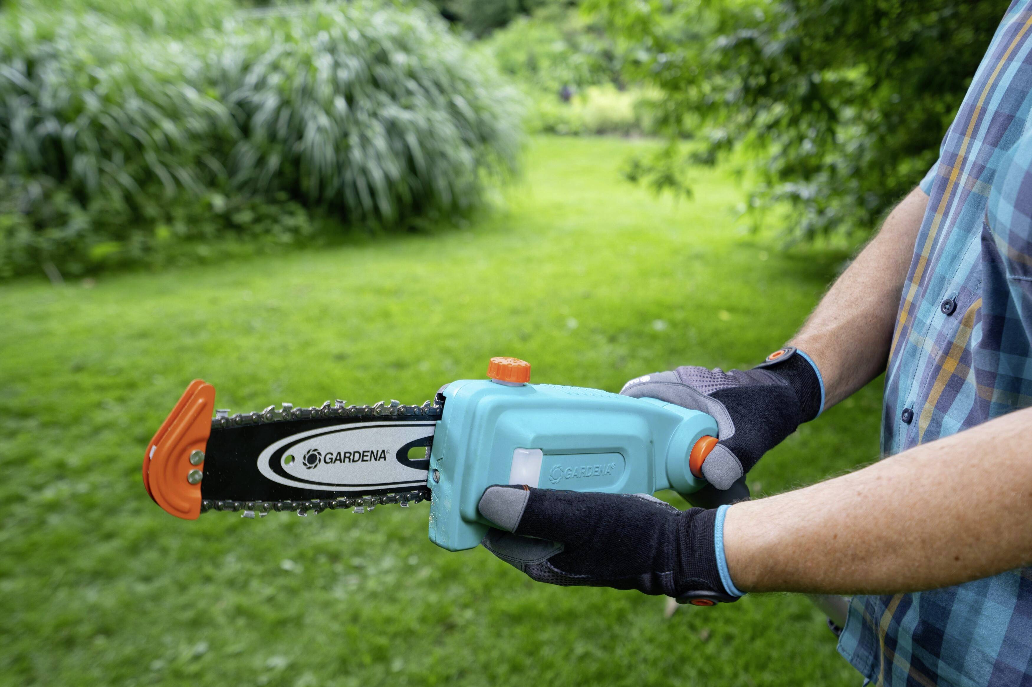 Eine Person hält eine blaue Spielzeug-Kettensäge im Garten. Die Säge hat eine orangefarbene Abdeckung und wird von behandschuhten Händen gehalten.