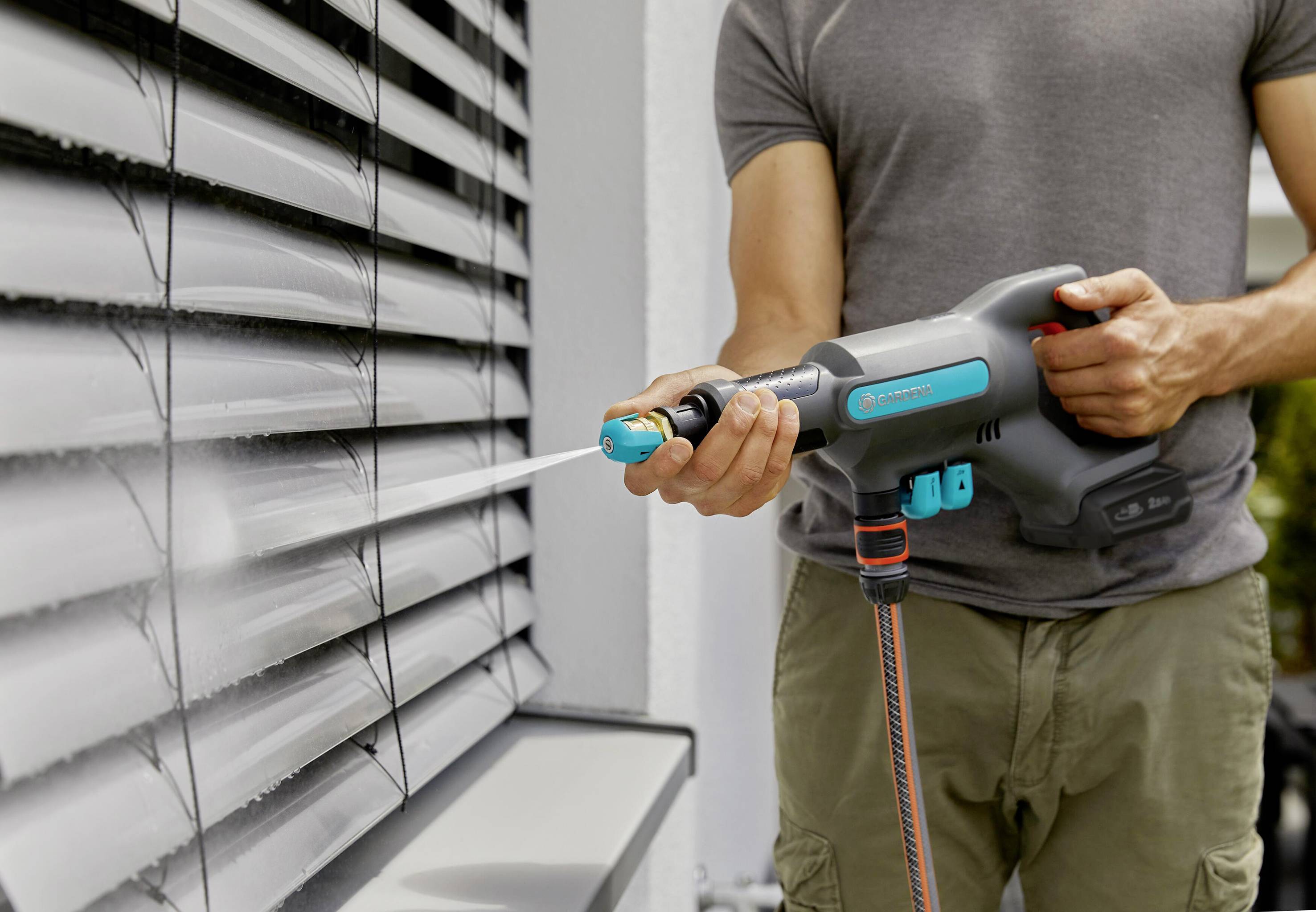 Eine Person reinigt mit einer elektrischen Gartenspritze die Jalousien eines Hauses, sprüht Wasser auf den Staub.
