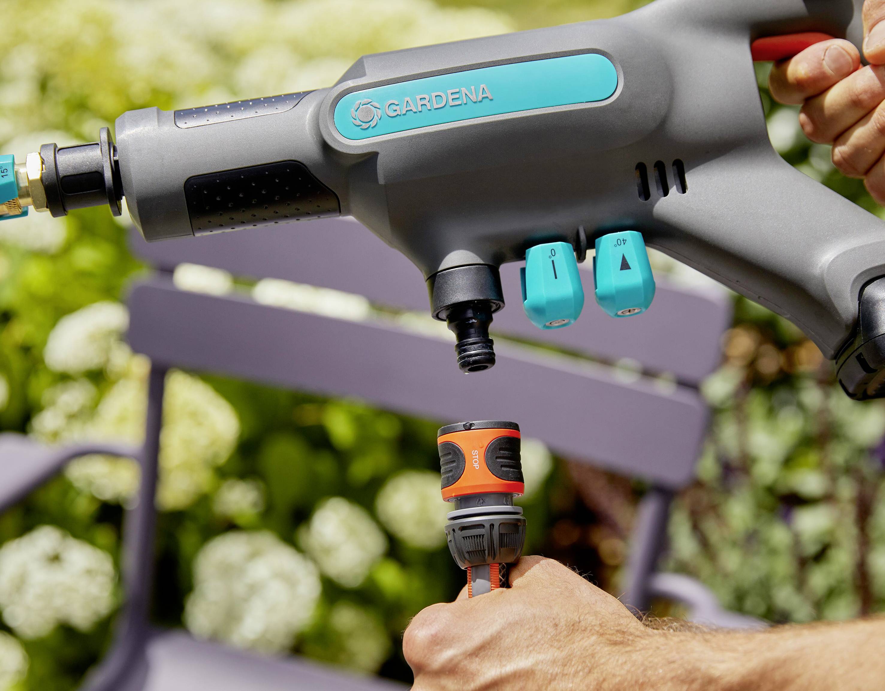 Eine Person verbindet einen GARDENA-Wasseranschluss mit einer Sprühpistole im Garten, Hintergrund zeigt Pflanzen und Gartenstuhl.
