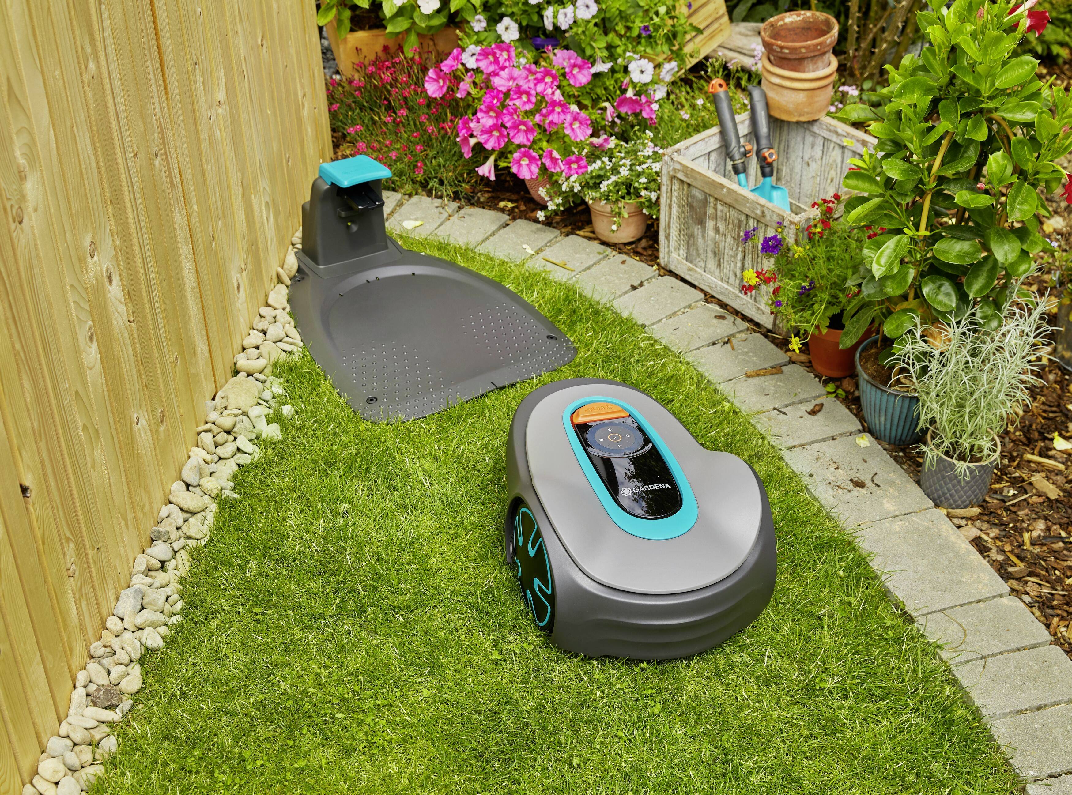 Ein Rasenmähroboter mäht den Rasen in einem Garten. Im Hintergrund blühen bunte Blumen und es steht ein Holzregal mit Gartenwerkzeugen.