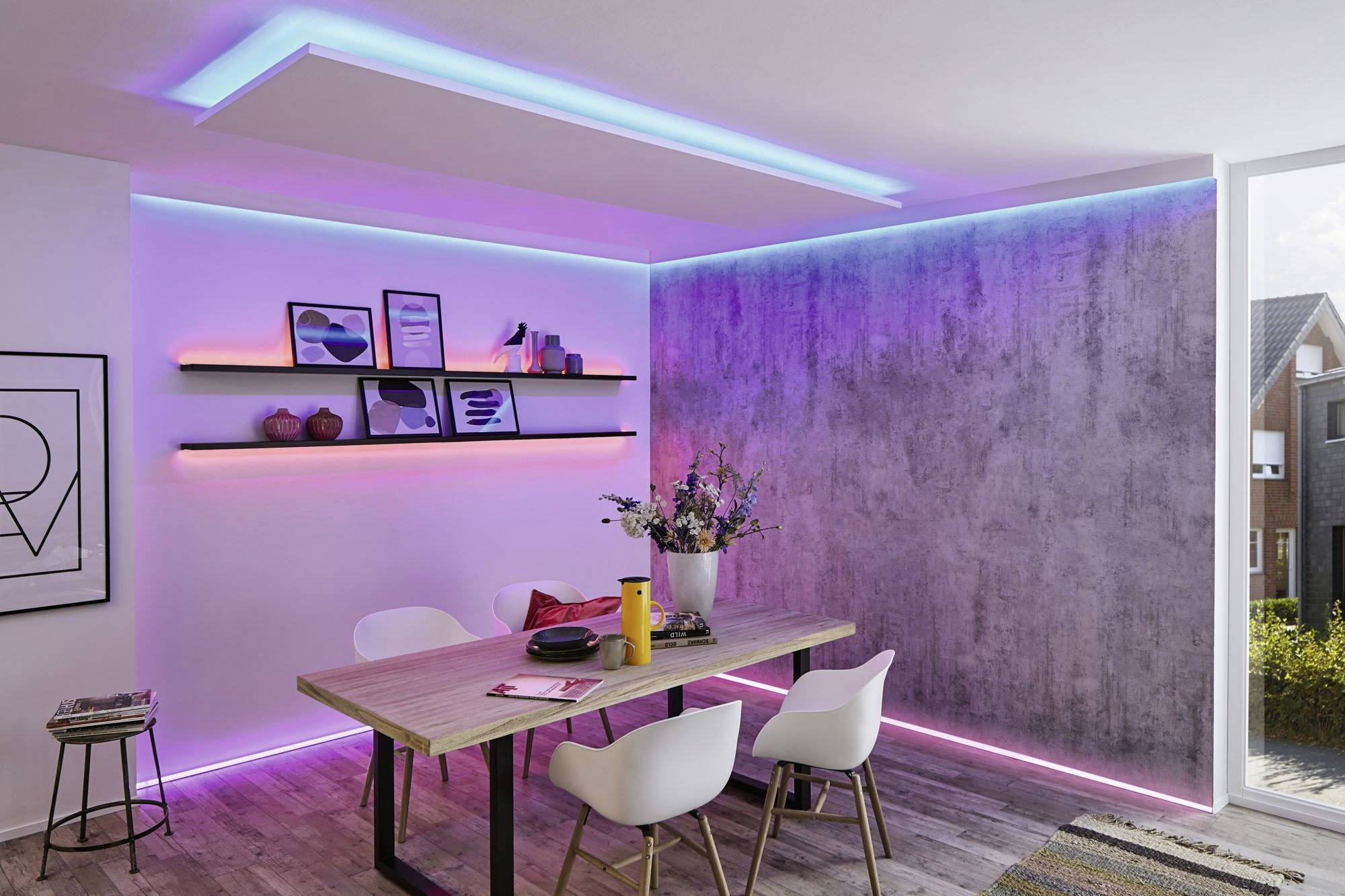 Esszimmer mit Holztisch, vier weißen Stühlen und farbiger LED-Beleuchtung an Wänden und Decke. Regale mit Dekor und Blumen auf Tisch.