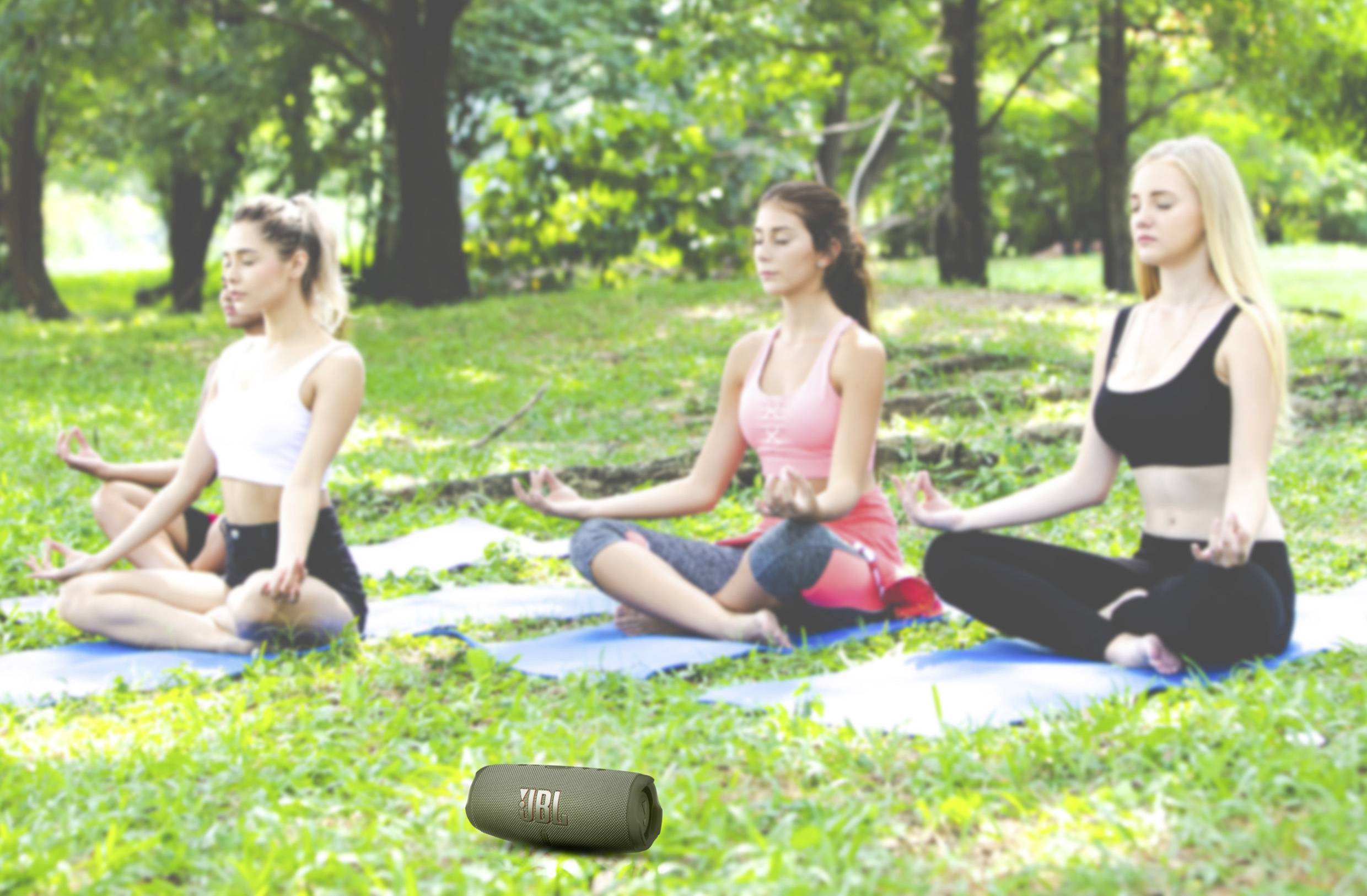 Drei Personen meditieren auf Yogamatten im Freien in einem grünen Park. Sie sitzen im Schneidersitz mit geschlossenen Augen.