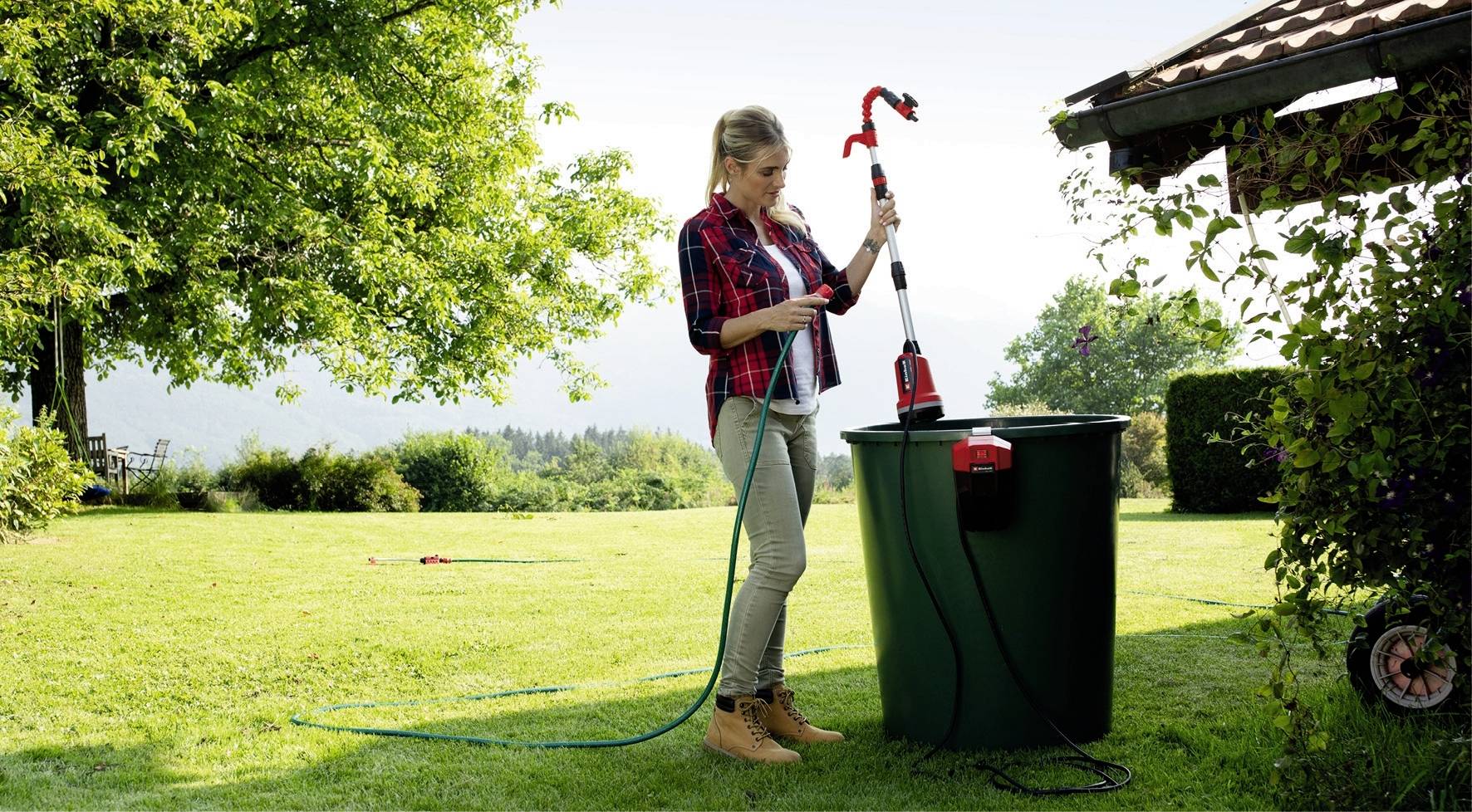 Eine Frau steht im Garten neben einem großen Wassertank und hält eine Pumpe. Sie legt den Schlauch in den Tank, um Wasser zu fördern.