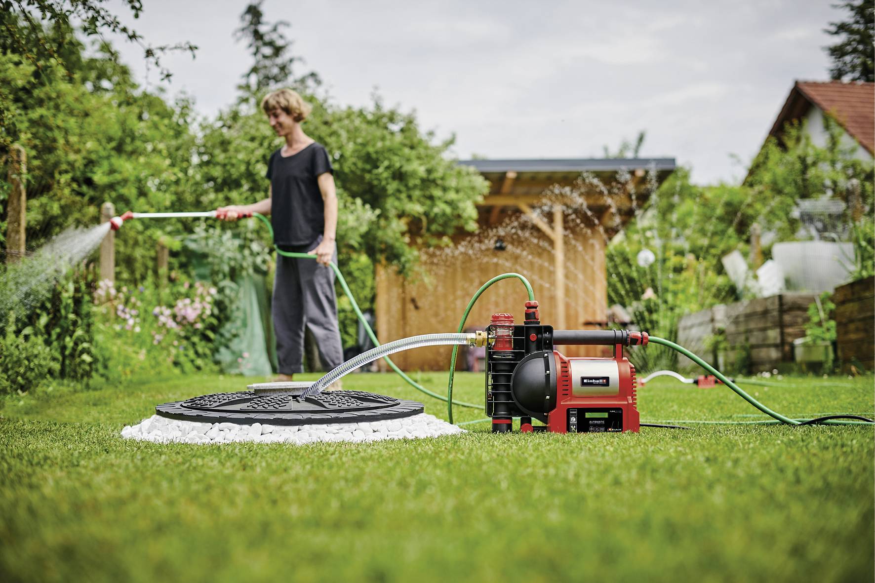 Person bewässert Garten mit einem Wasserschlauch. Im Vordergrund eine rote Wasserpumpe, die an einen unterirdischen Tank angeschlossen ist.