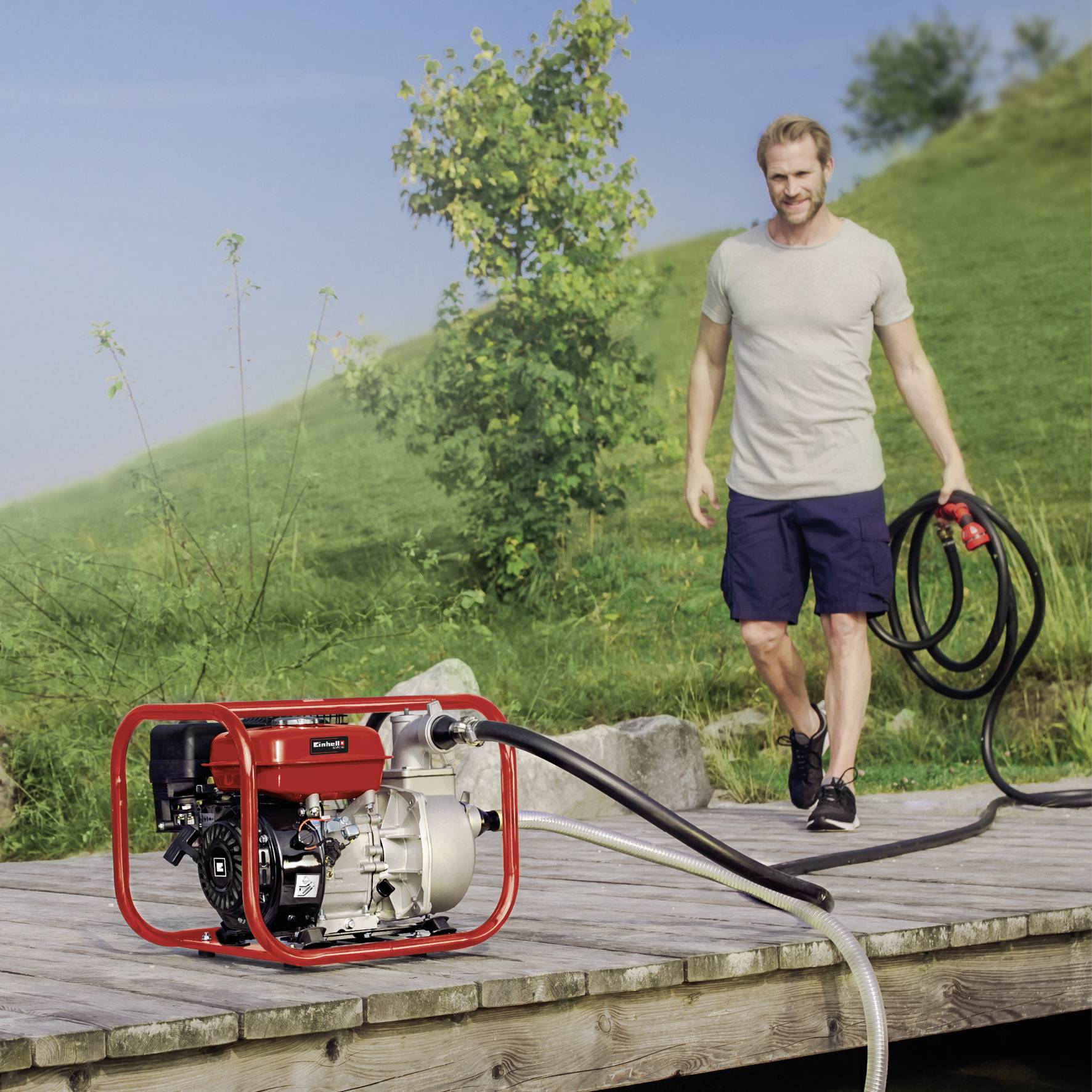 Ein Mann auf einem Steg hält einen Wasserschlauch, der mit einem roten Wasserpumpe am Boden verbunden ist. Im Hintergrund ist Gras und ein Baum.