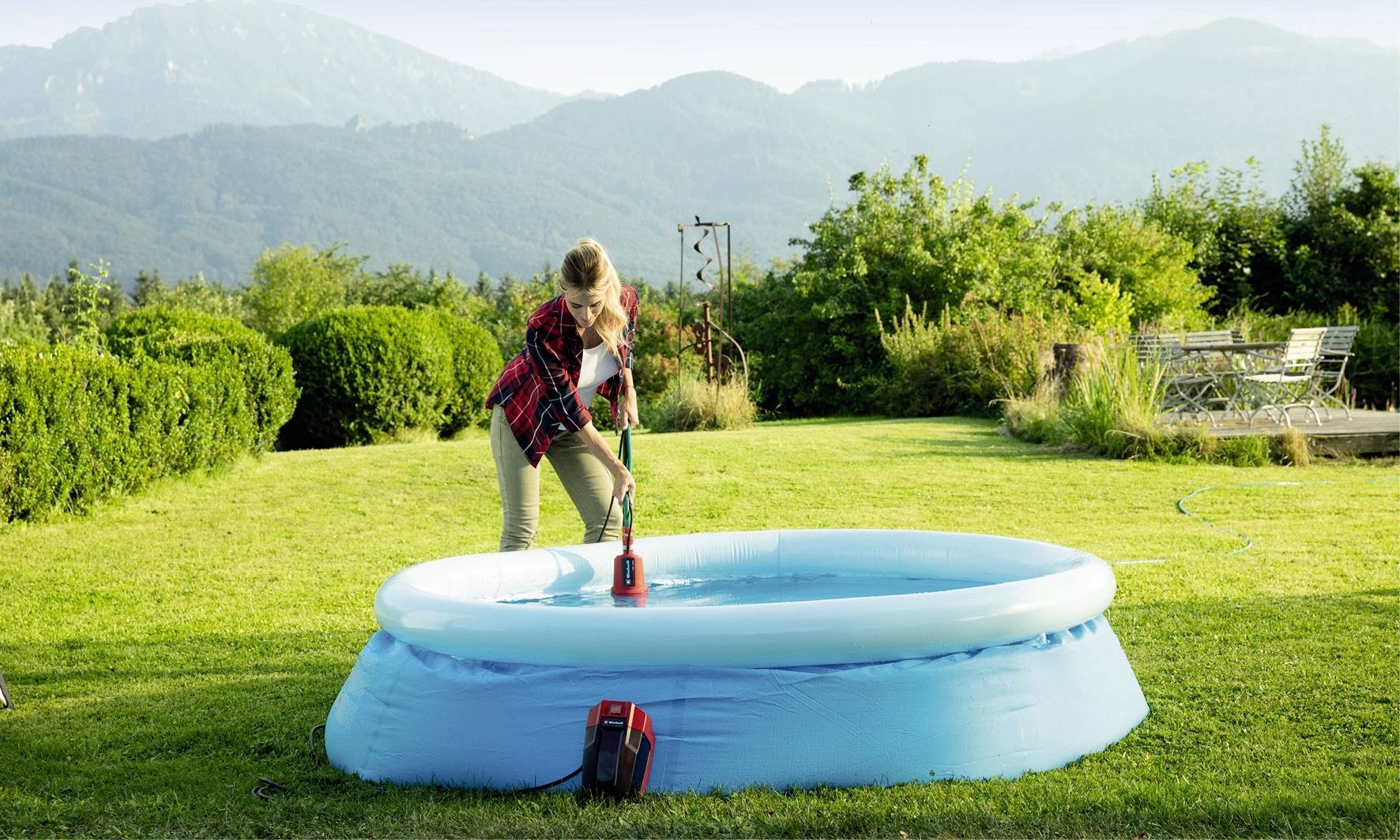 Eine Person reinigt ein aufblasbares Planschbecken mit einer Bürste in einem Garten mit Bergblick im Hintergrund.