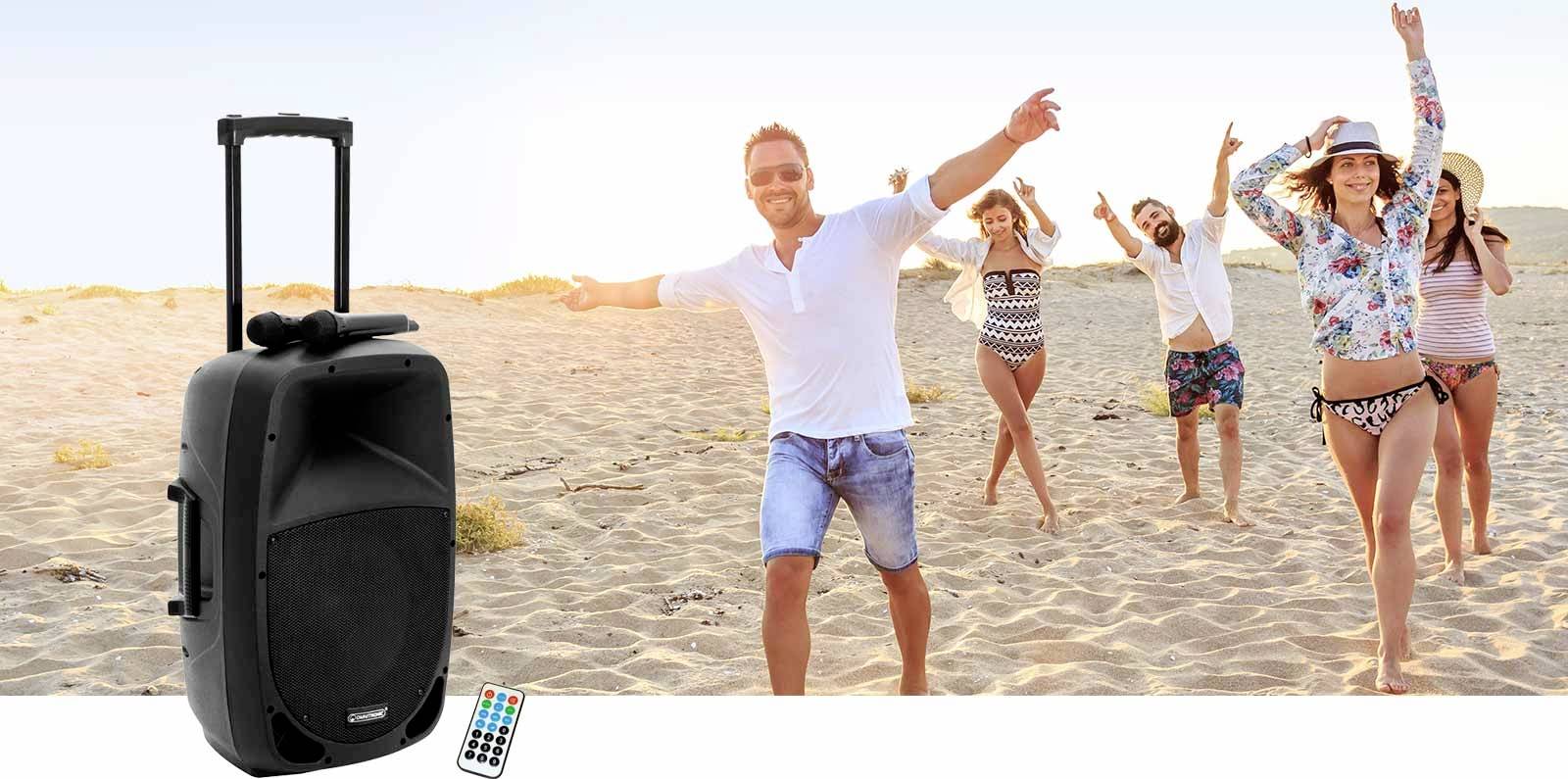 Eine Gruppe junger Erwachsener tanzt fröhlich am Strand bei Sonnenuntergang. Links im Bild befindet sich ein tragbarer Lautsprecher mit Fernbedienung.