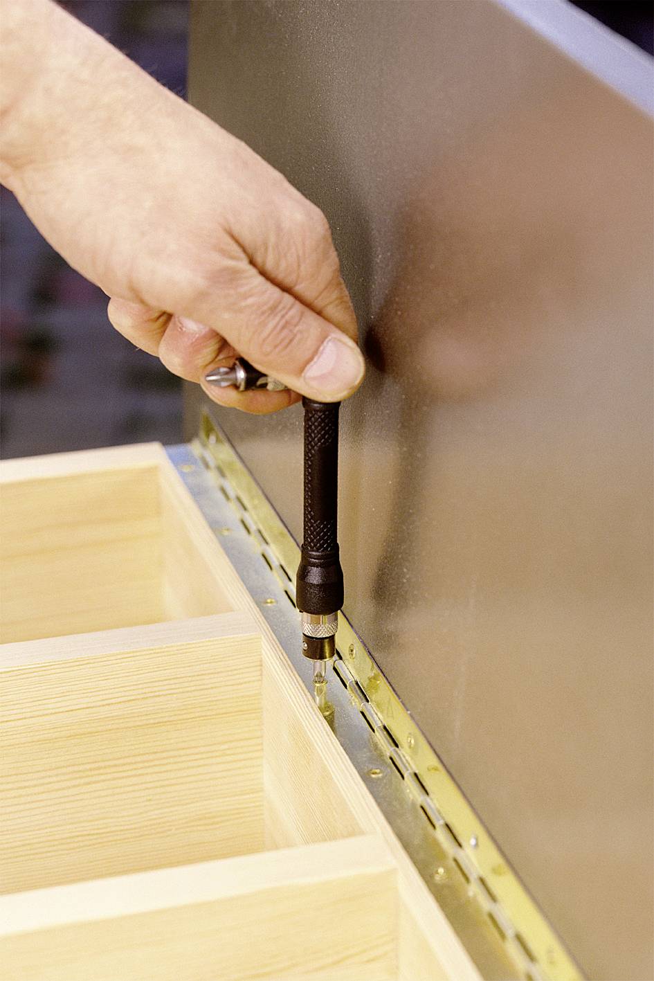 Eine Hand befestigt mit einem Schraubendreher ein Scharnier an der Kante eines Holzstücks, möglicherweise für ein Möbelstück.