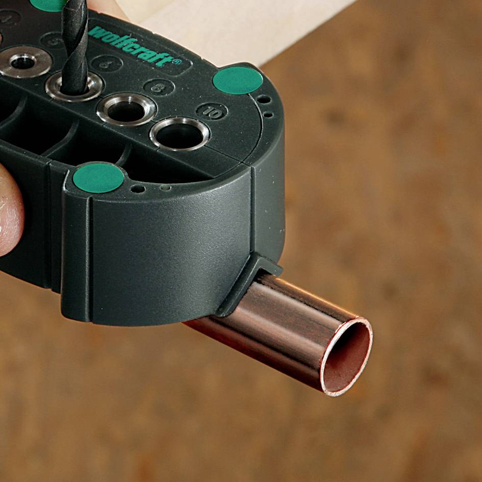 Eine Hand hält ein Werkzeug, das ein Loch in ein Kupferrohr bohrt. Das Werkzeug hat eine grüne Oberfläche mit Markierungen.