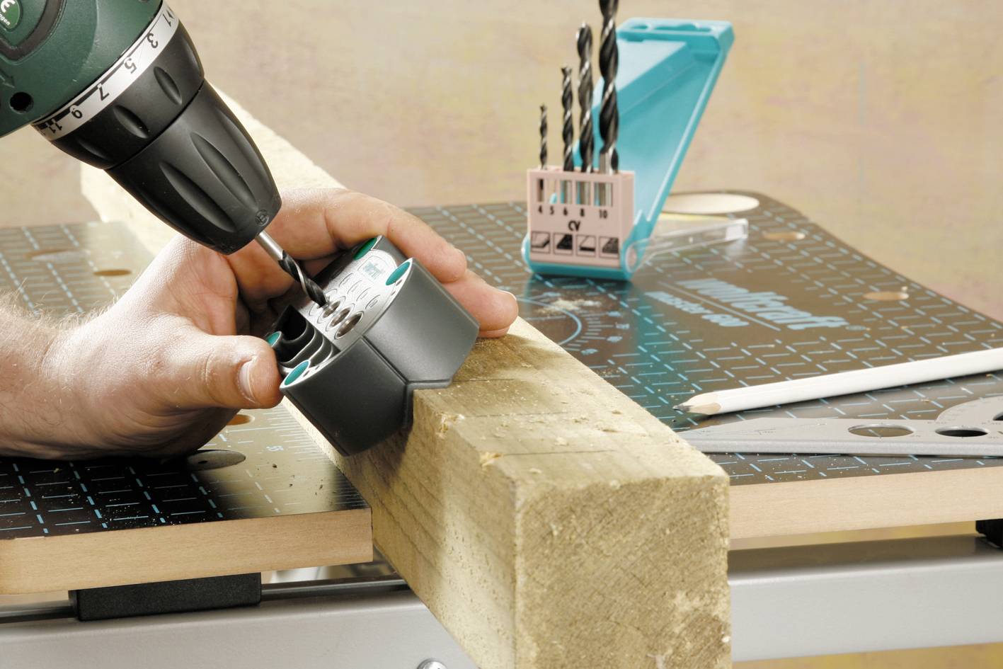 Eine Person bohrt mit einer grünen Bohrmaschine Löcher in einen Holzblock, während eine Schachtel mit Bohrern und ein Bleistift daneben liegen.