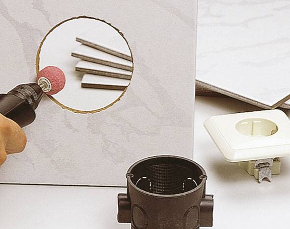 Ein Handwerker bearbeitet ein rundes Loch in einer Fliese mit einem Schleifwerkzeug. Im Vordergrund liegen Installationsmaterialien.