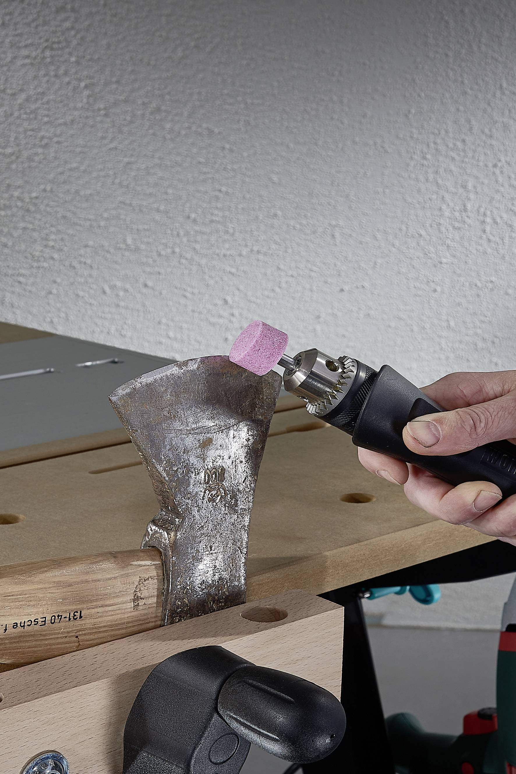 Eine Person schleift die stumpfe Kante einer Axt mit einem rotierenden Schleifwerkzeug ab. Die Axt ist in einem Schraubstock eingespannt.