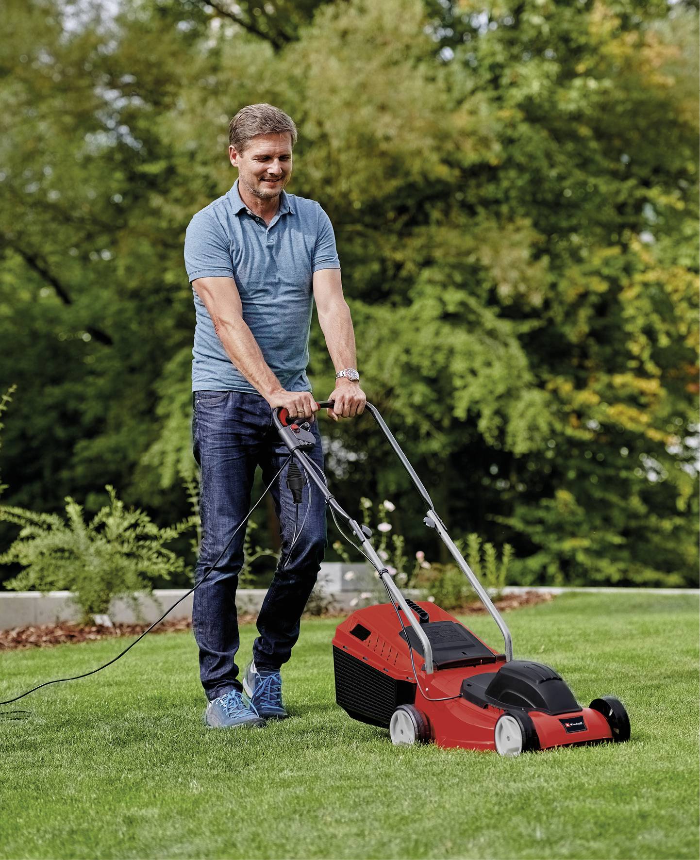 Ein Mann mäht den Rasen in einem Garten mit einem roten Elektrorasenmäher. Im Hintergrund sind Bäume und Büsche zu sehen.