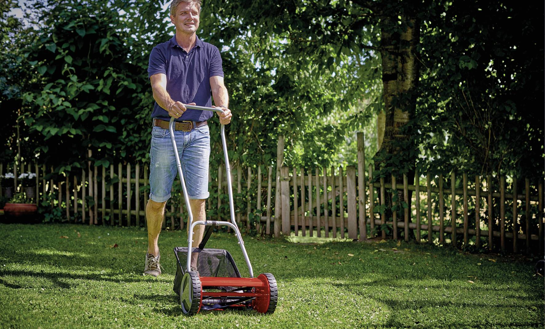 Person im blauen Shirt und Jeansshorts mäht mit einem handbetriebenen Rasenmäher den Rasen in einem grünen Garten.