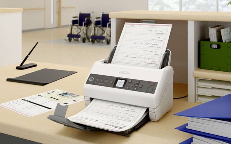 Ein Scanner scannt Dokumente in einem Büro. Auf dem Tisch liegen blaue Ordner und ein schwarzes Klemmbrett. Im Hintergrund stehen Rollstühle.