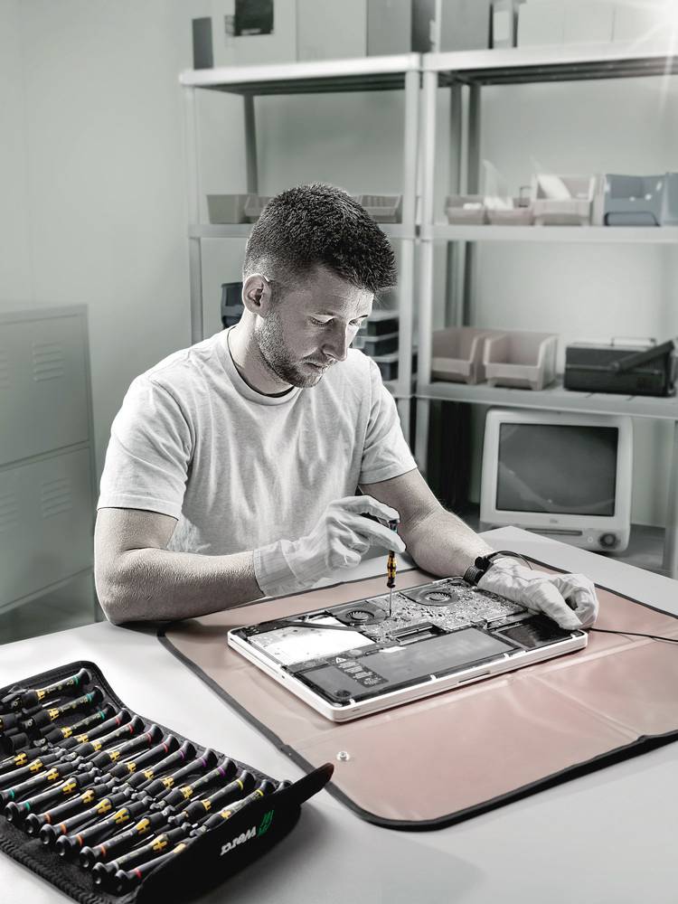 Ein Mann repariert ein elektronisches Gerät auf einem Tisch. Um ihn herum liegen Werkzeuge. Im Hintergrund sind Regale zu sehen.