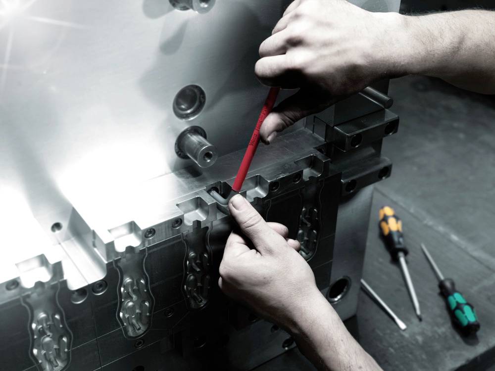 Jemand justiert mit einem roten Schraubendreher eine Metallmaschine. Im Hintergrund liegen zwei weitere Schraubendreher.