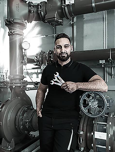 Eine Person in Arbeitskleidung mit Schraubenschlüsseln steht in einer industriellen Umgebung mit Rohren und Maschinen, was auf Wartungsarbeiten hindeutet.