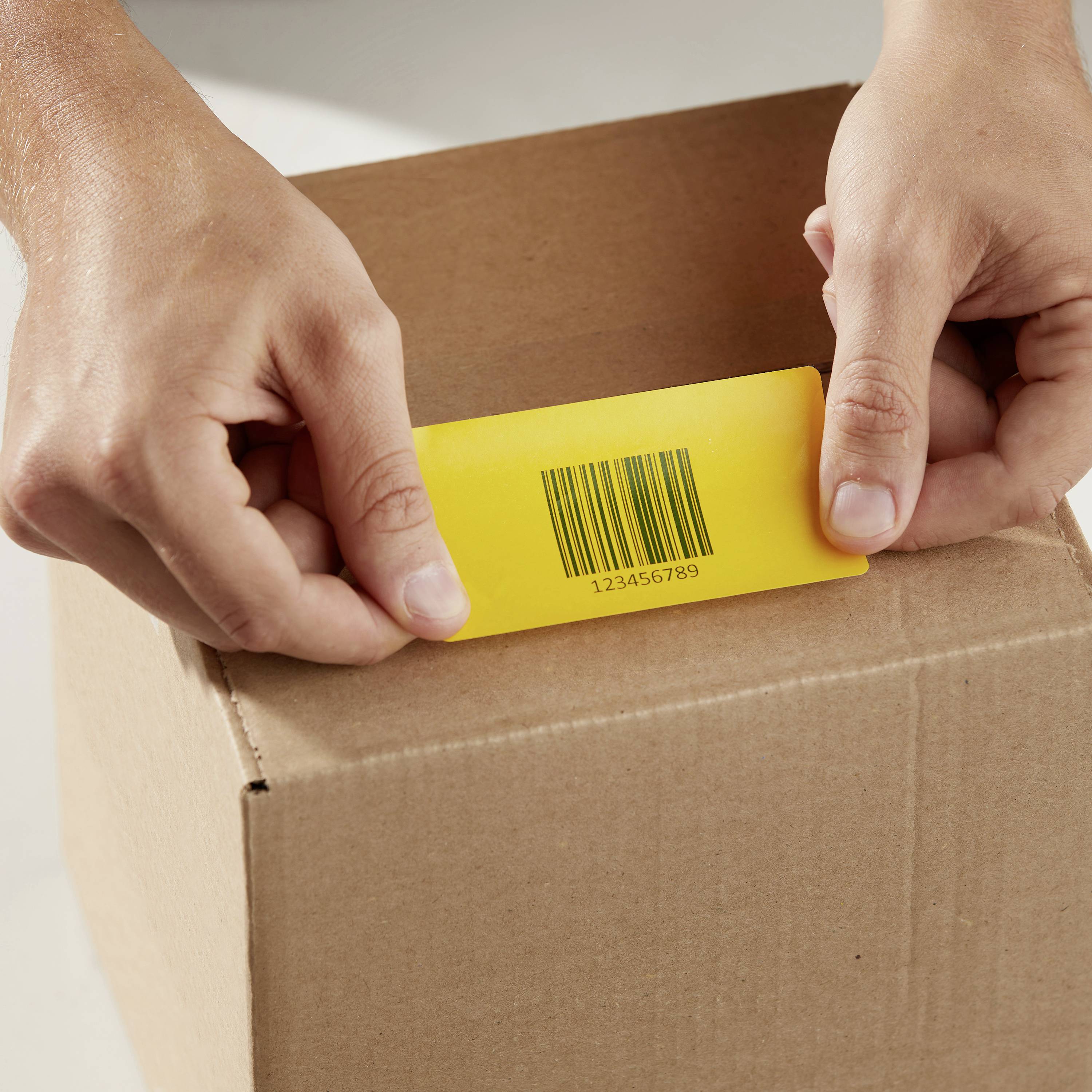 Hände halten eine gelbe Karte mit einem Barcode '123456789', die in einen braunen Karton gesteckt wird.
