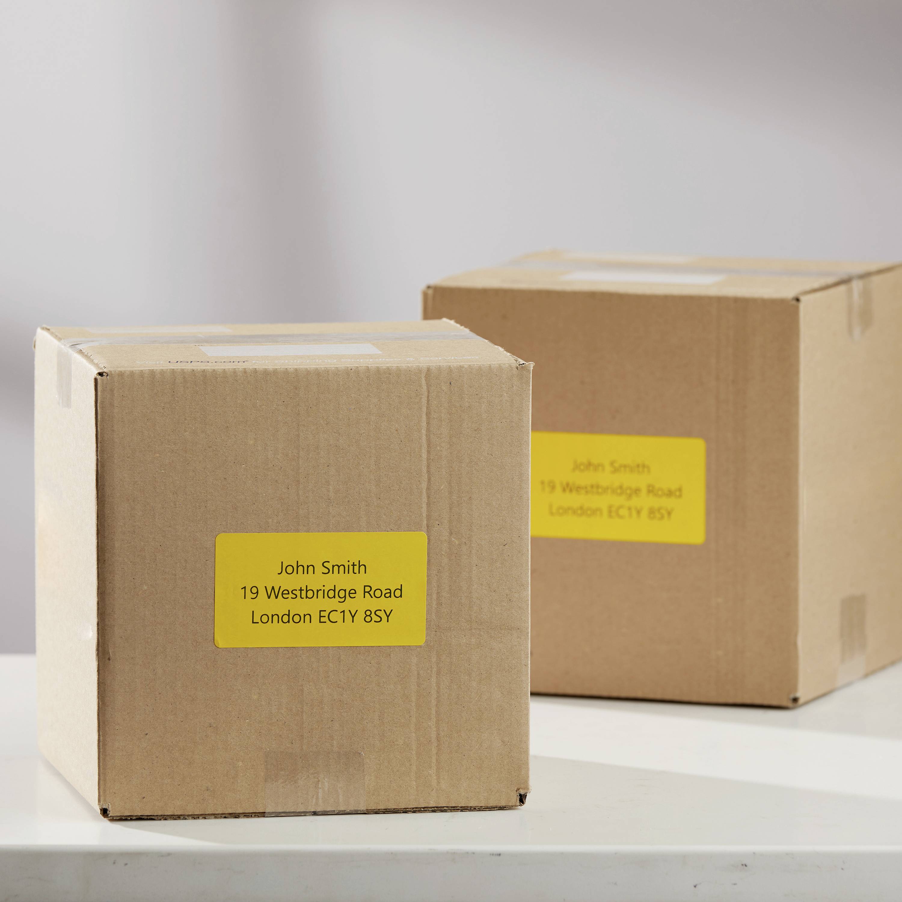 Zwei Pakete auf einem Tisch, beide mit gelbem Etikett. Aufschrift: 'John Smith, 19 Westridge Road, London EC1Y 8SY'.