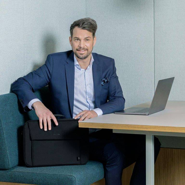 Ein Mann im Anzug sitzt in einem Bürobereich mit einem Laptop, Blick zur Kamera, mit einer Mappe auf dem Tisch.