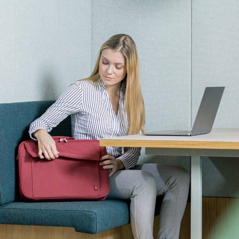 Eine Frau sitzt an einem Tisch und öffnet eine rote Tasche. Neben ihr steht ein Laptop. Sie trägt eine gestreifte Bluse.