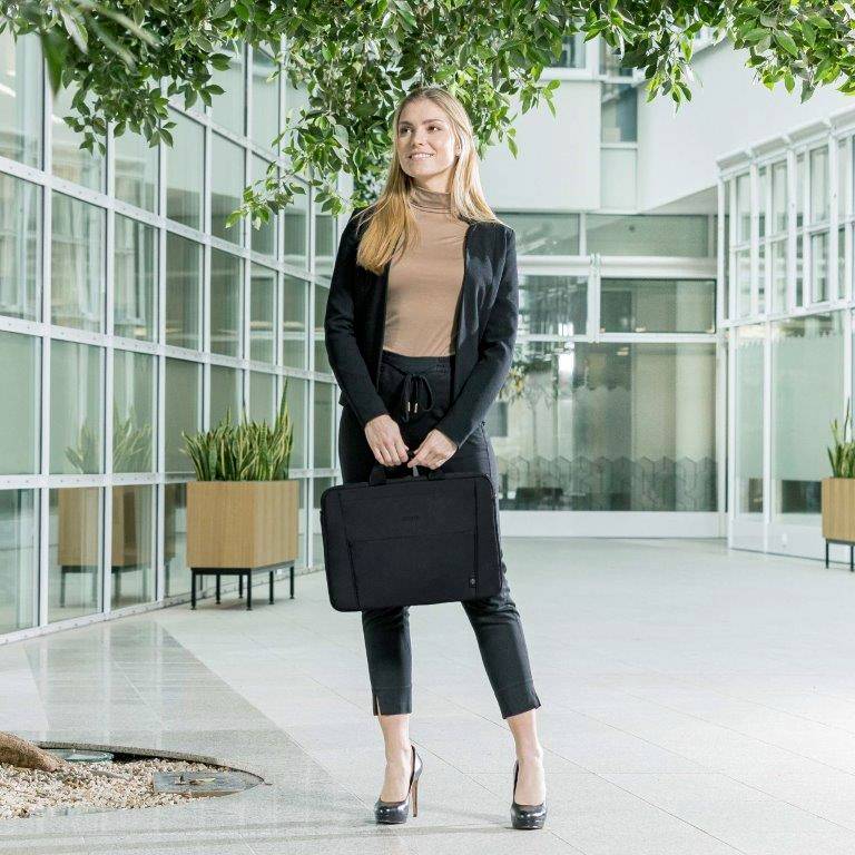 Frau steht in modernem Bürogebäude, trägt schwarze Kleidung, hält eine schwarze Aktentasche. Große Fenster und Pflanzen im Hintergrund.