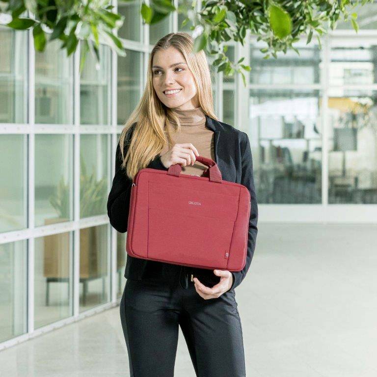 Eine Frau steht in einem modernen Büro mit einem roten Laptop-Case in der rechten Hand, umgeben von Pflanzen und Glaswänden.
