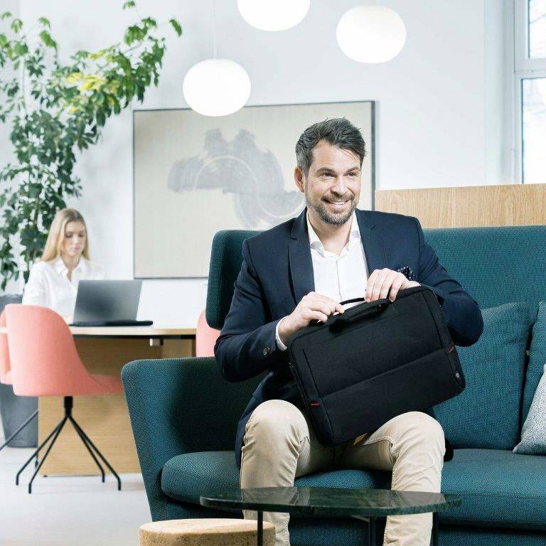 Ein Mann im Anzug sitzt auf einem Sofa und hält eine Tasche; im Hintergrund arbeitet eine Frau am Laptop an einem Schreibtisch.
