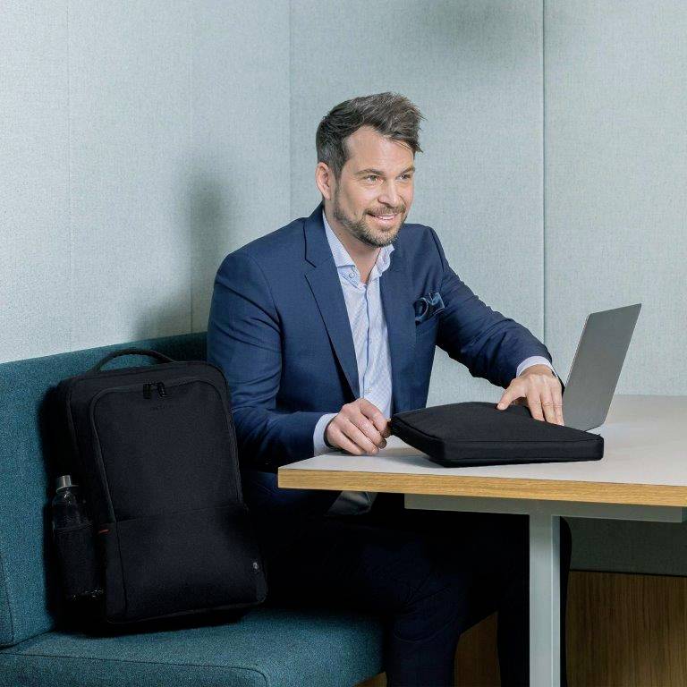 Ein Mann in einem blauen Anzug sitzt in einem modernen Bürobereich mit einem Tisch und einem schwarzen Rucksack. Er sieht freundlich aus.