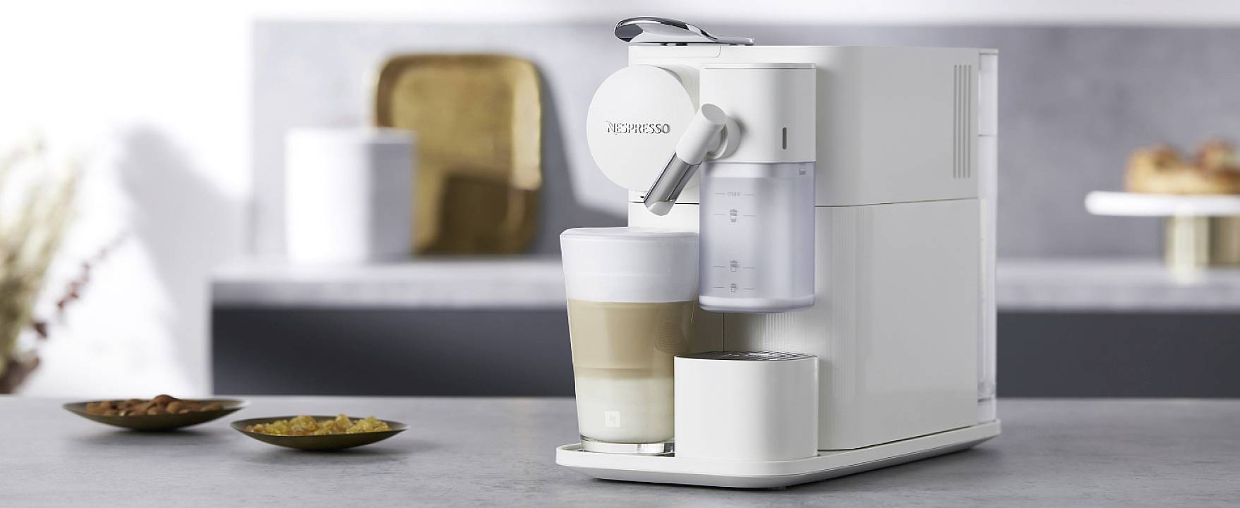 Weiße Kaffeemaschine auf grauer Arbeitsplatte, bereitet einen Cappuccino zu. Im Hintergrund unscharfe Küchelemente und Teller.