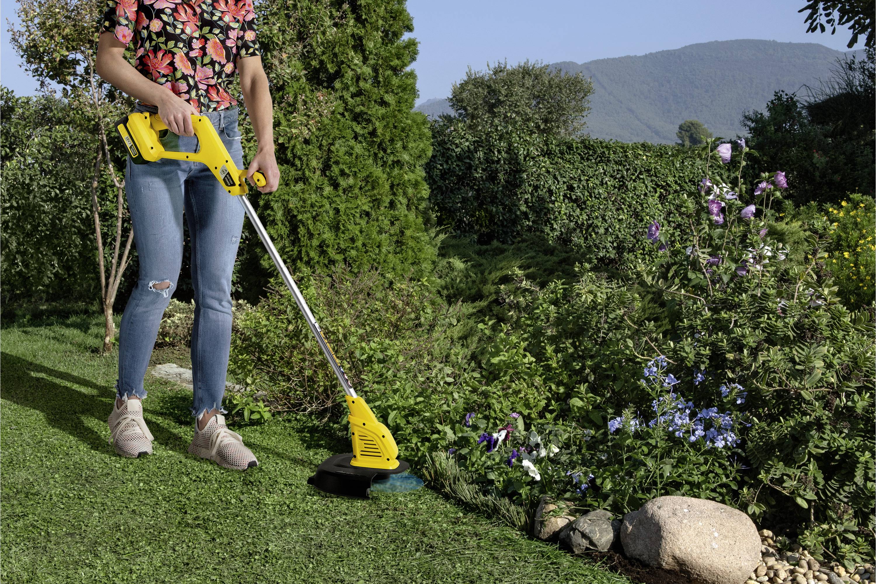 Person nutzt eine elektrische Rasentrimmer, um einen gepflegten Garten mit bunten Blumen und Büschen nahe einem Berg zu schneiden.