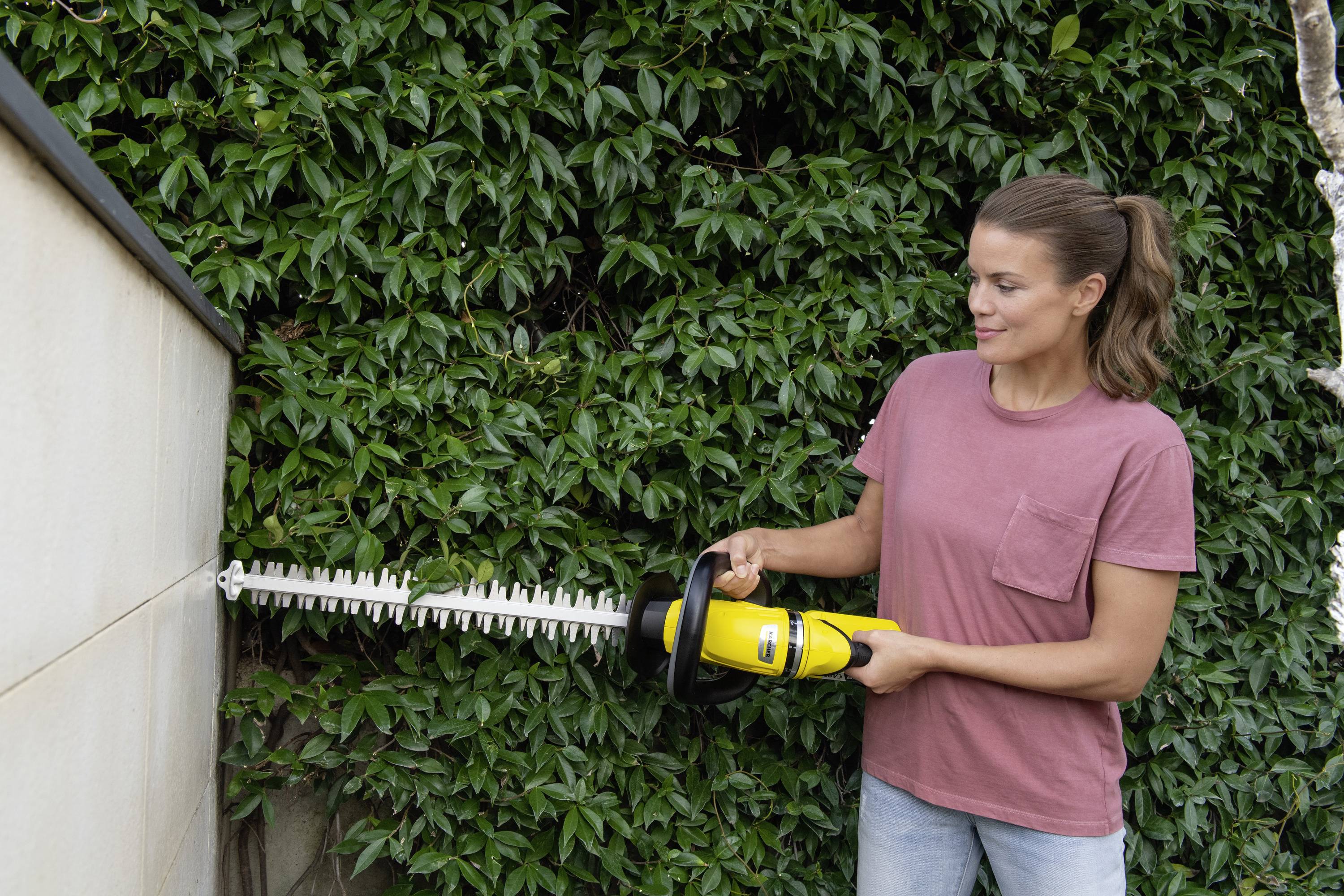 Eine Frau in einem rosa T-Shirt schneidet mit einer Heckenschere eine dichte Hecke. Der Hintergrund ist eine grüne Wand aus Blättern.