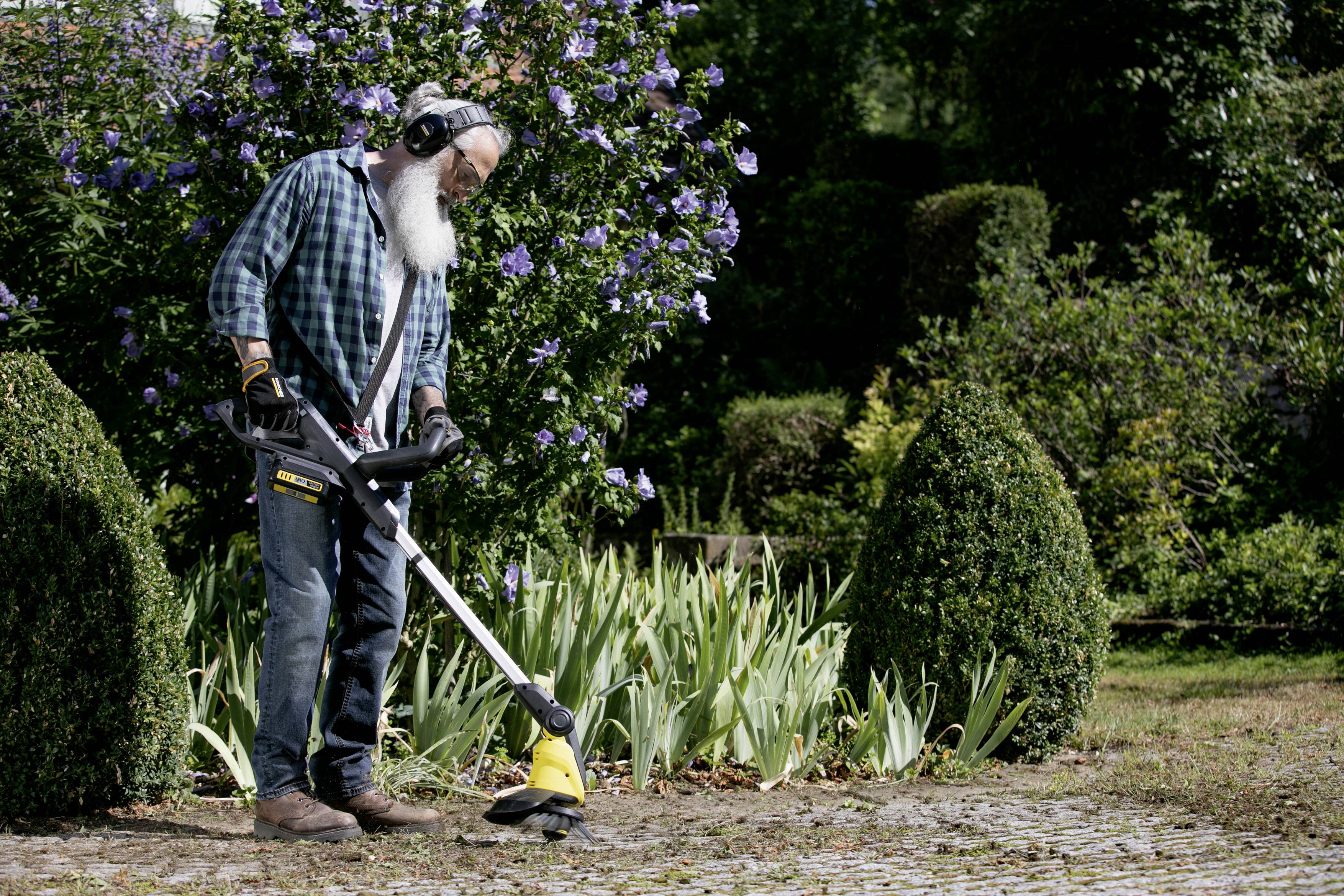 Eine Person benutzt im Garten einen Rasentrimmer, um Gehwege freizuhalten, umgeben von Blumen und Sträuchern. Sie trägt Gehörschutz.