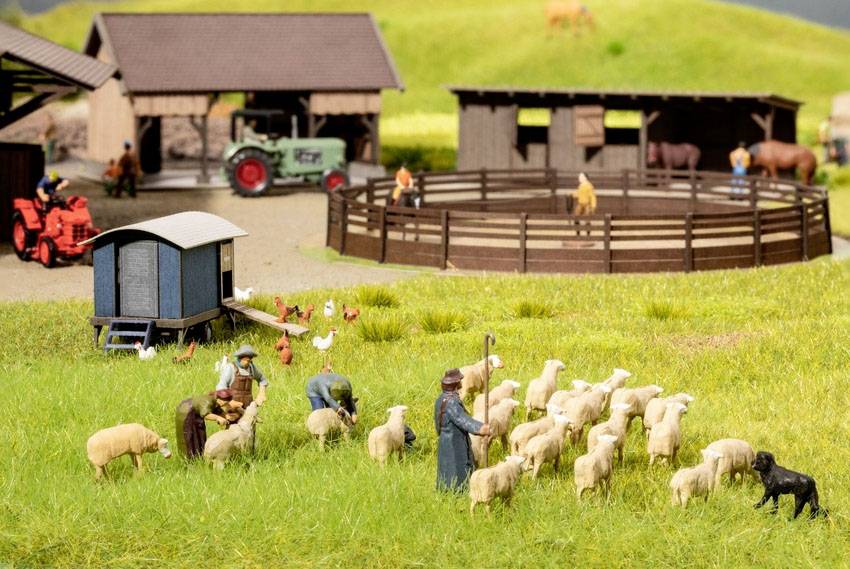Eine Miniatur-Farm mit Figuren, die Schafe hüten, umgeben von Hütten, Traktoren und Tieren. Ländliche Szene mit Menschen und Tieren im Vordergrund.