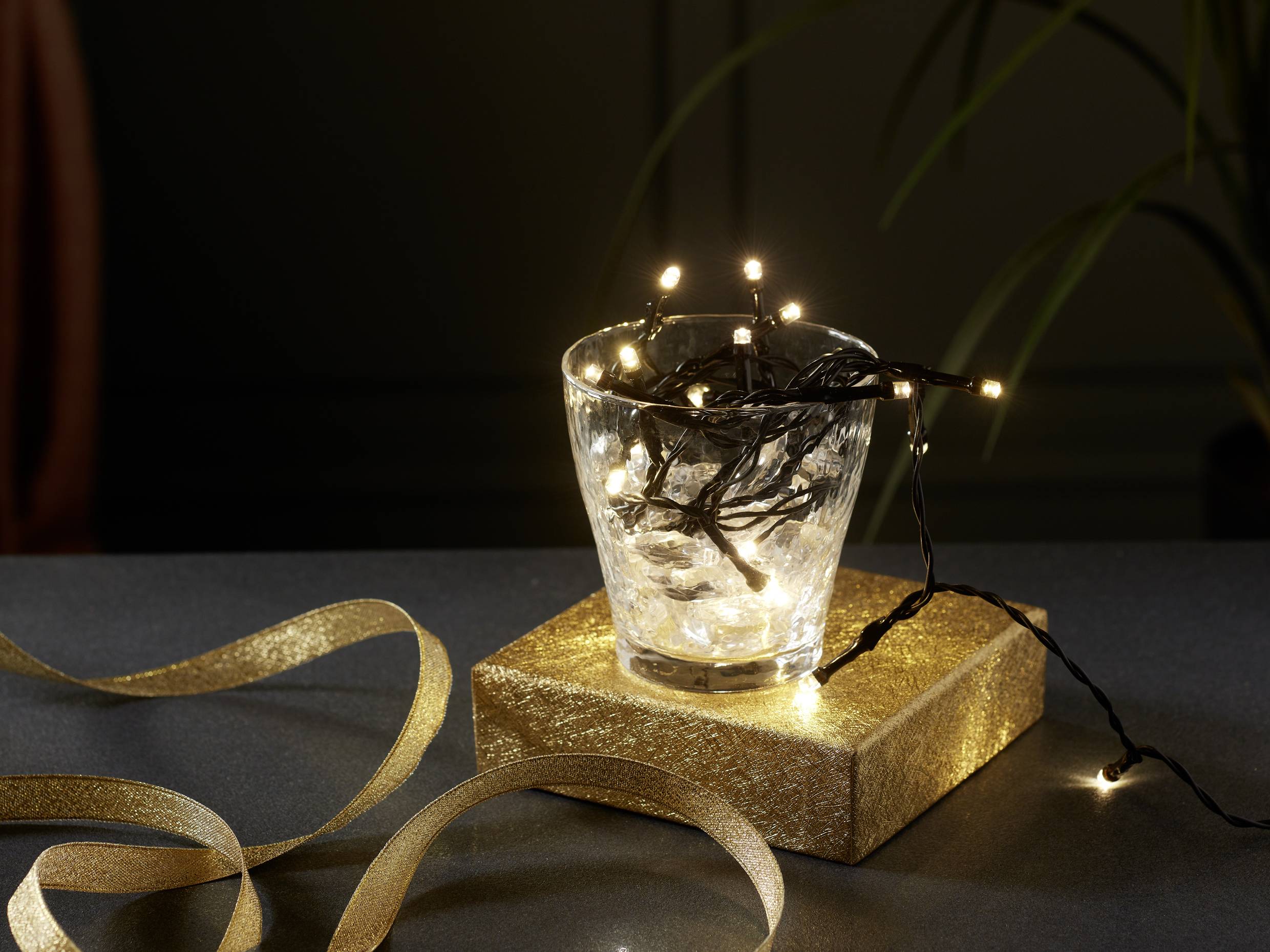 Ein Glas mit leuchtender Lichterkette steht auf einer goldenen Box auf einem Tisch, umgeben von goldfarbenen Bändern.