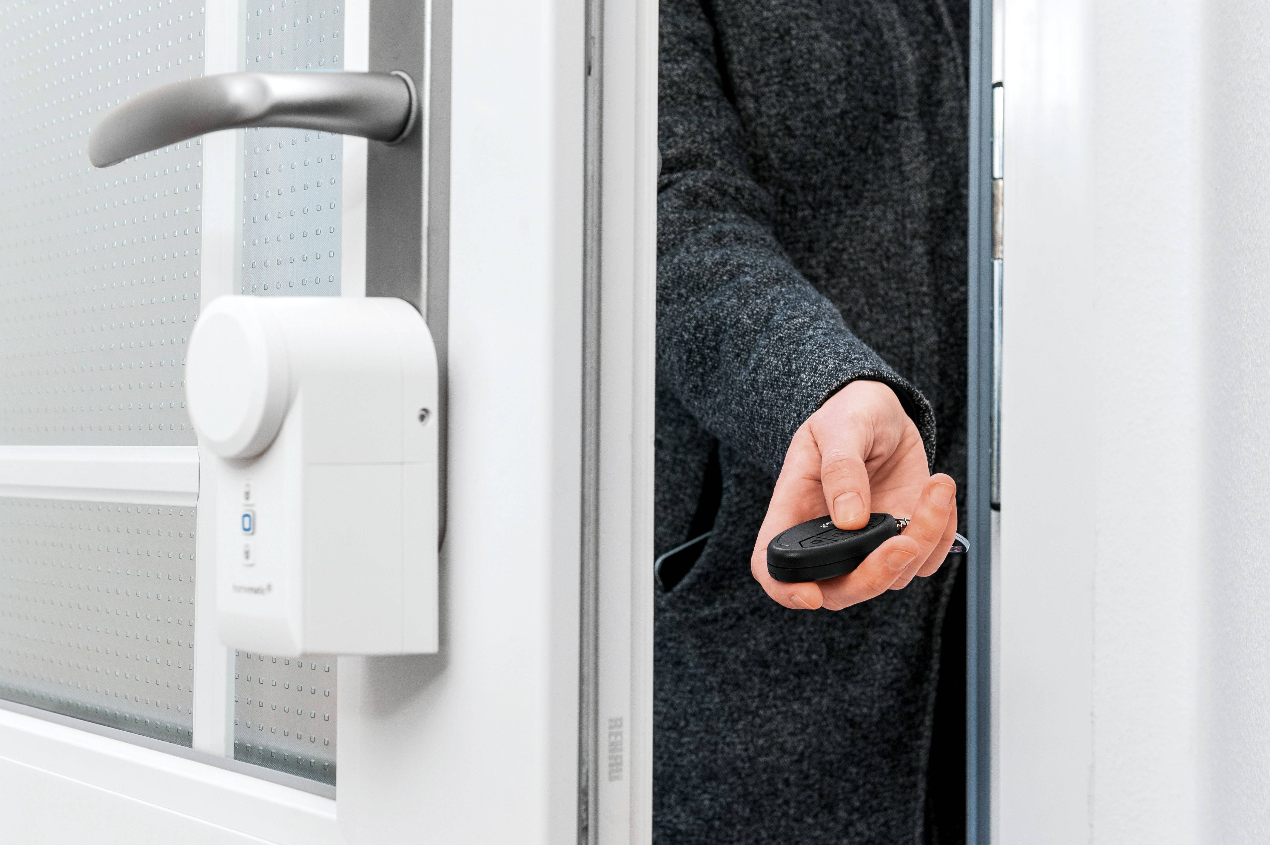 Eine Person in einem Mantel öffnet eine Tür mit einem elektronischen Türschloss, hält dabei einen Fernbedienung.