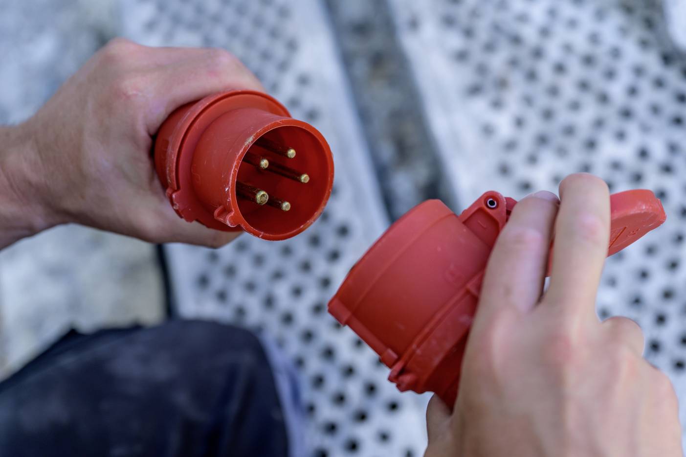 Zwei Hände verbinden einen roten, industriellen Stecker mit einer passenden Steckdose. Hintergrund unscharf, Fokus auf Steckverbindung.